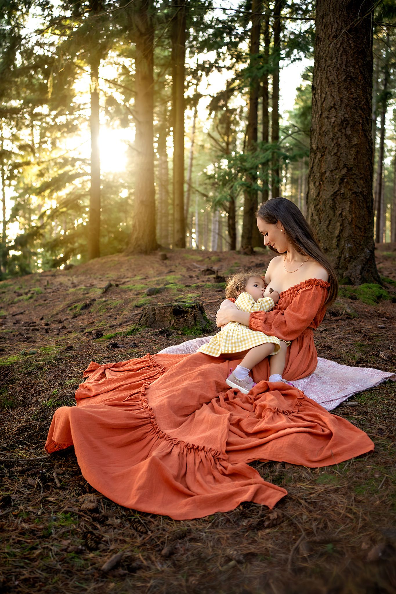 Mother in a beautiful gown, breastfeeding her child, sun looking through the trees in the forest. Location: New Forest, Hampshire.