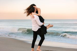 Two silhouettes embrace and dance playfully on a sandy beach at sunset with crashing ocean waves in the background.