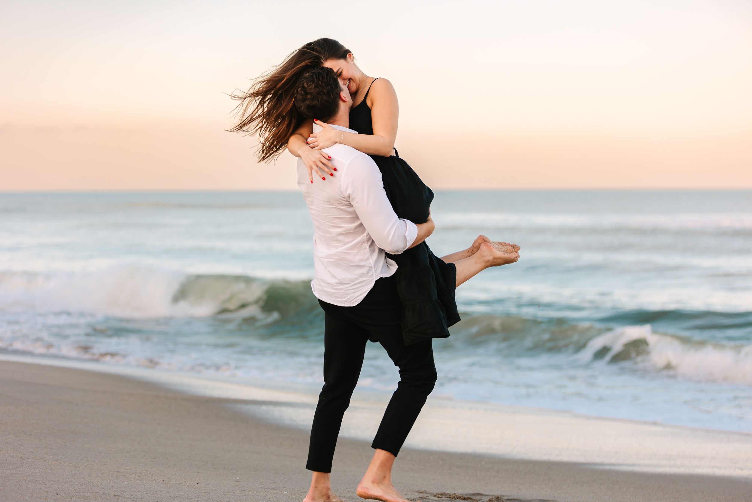 Two silhouettes embrace and dance playfully on a sandy beach at sunset with crashing ocean waves in the background.