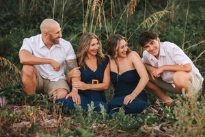 A group of friends laugh together while sitting in tall grass during a casual outdoor photoshoot at sunset.