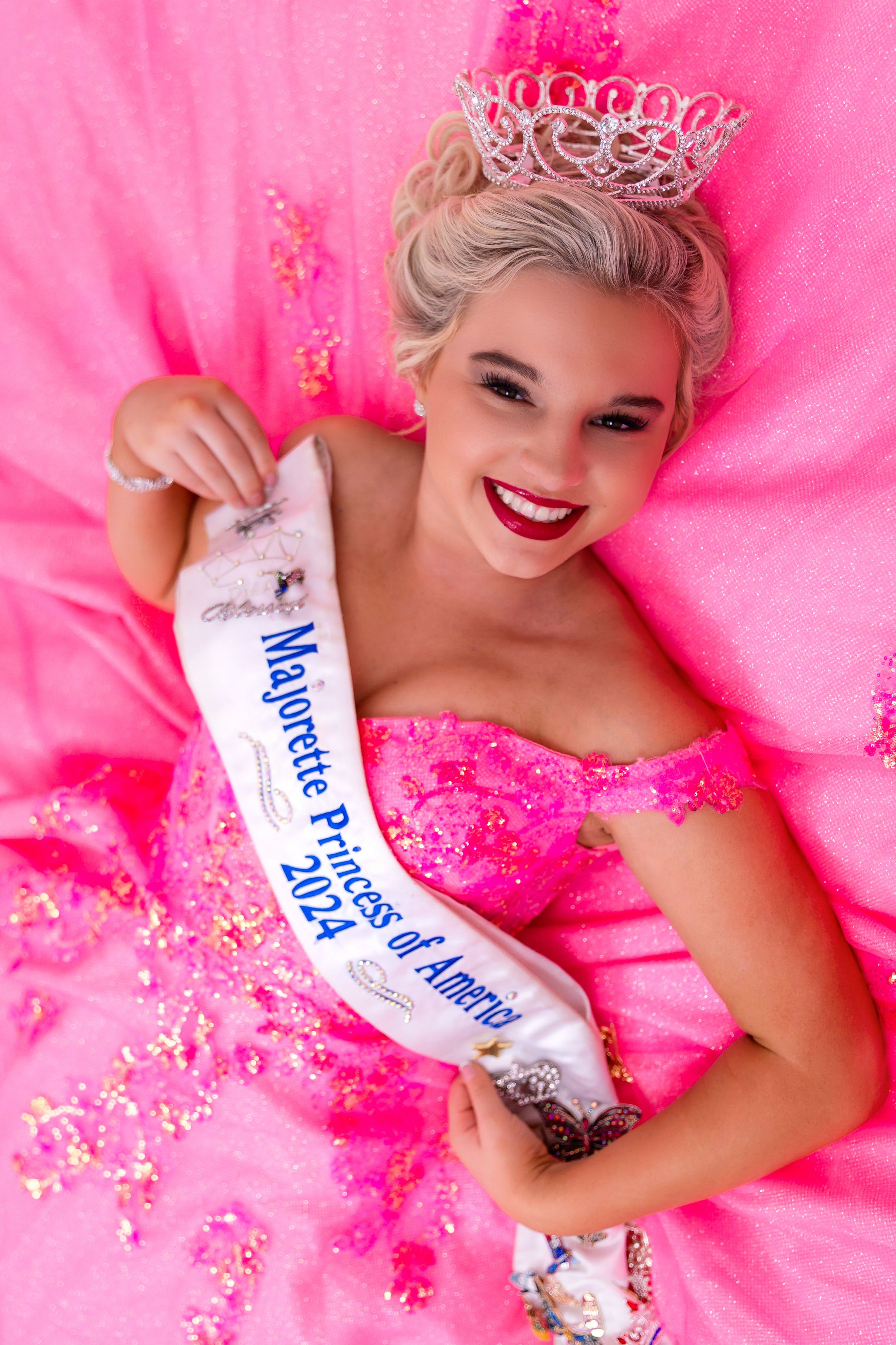 A beauty pageant contestant wearing a sparkling crown and pink gown poses with a sash against a bright pink background.