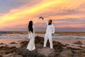 A couple in white outfits dances on coastal rocks during a dramatic orange sunset over the ocean.