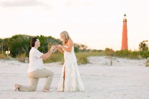A romantic proposal scene on a sandy beach with a red lighthouse in the background during sunset.