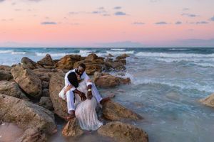 A couple in white attire shares an intimate moment on rocky coastal cliffs during a pink sunset.