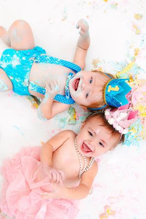Babies in colorful tutus lie on white backdrop with splashes of pink and blue paint creating artistic first birthday portraits.