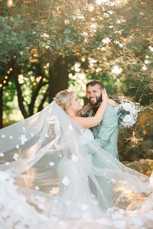 A couple in wedding attire dances outdoors with a flowing white veil creating ethereal movement in the sunlit garden.