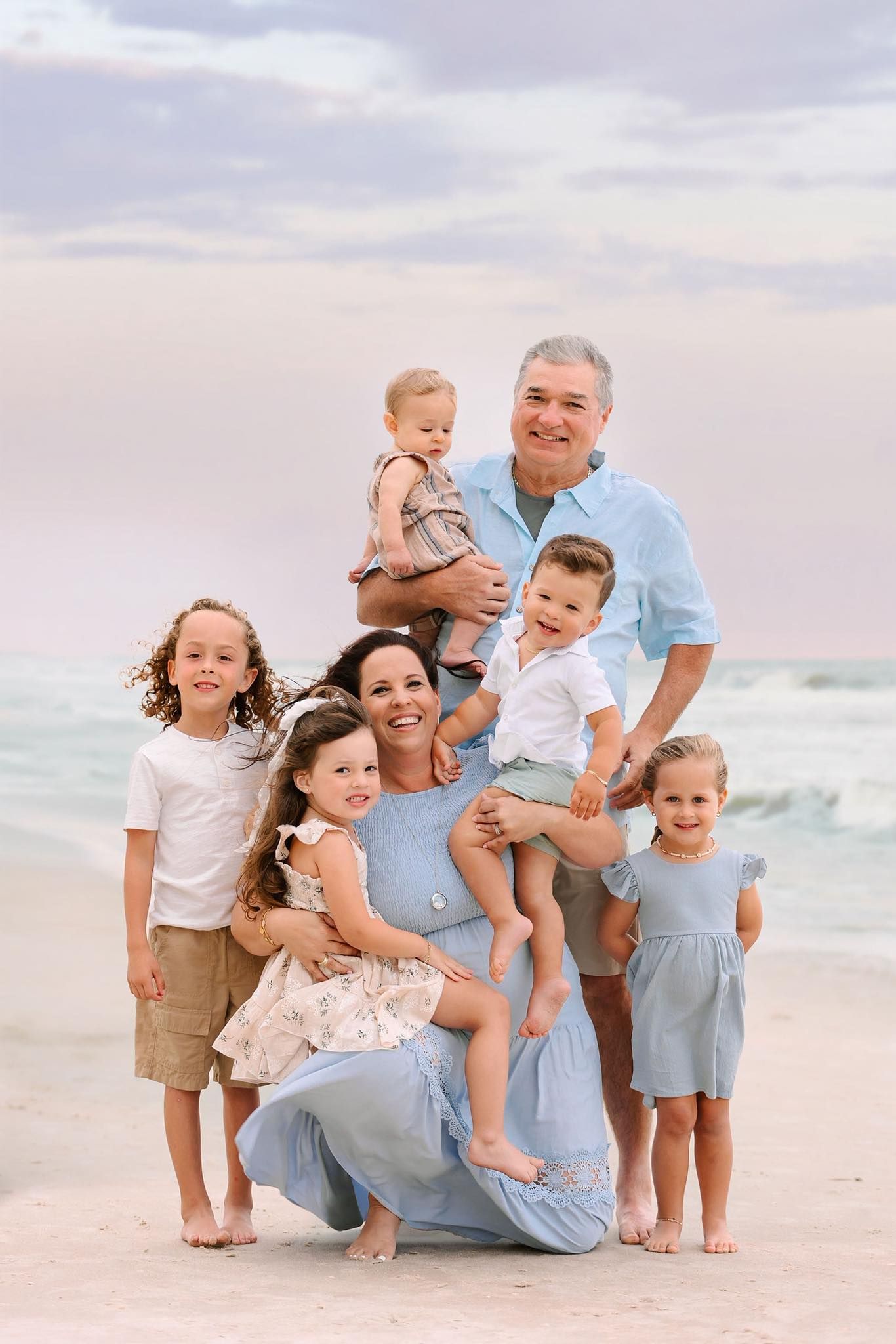 A happy family of seven poses together on a beach at sunset wearing coordinating blue and neutral colored outfits.