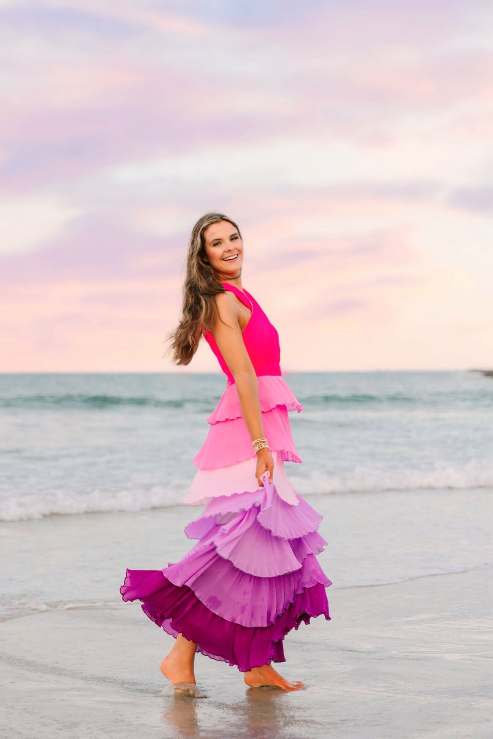 A woman in pink and purple ruffled dress twirls joyfully on the beach at sunset with ocean waves in background.