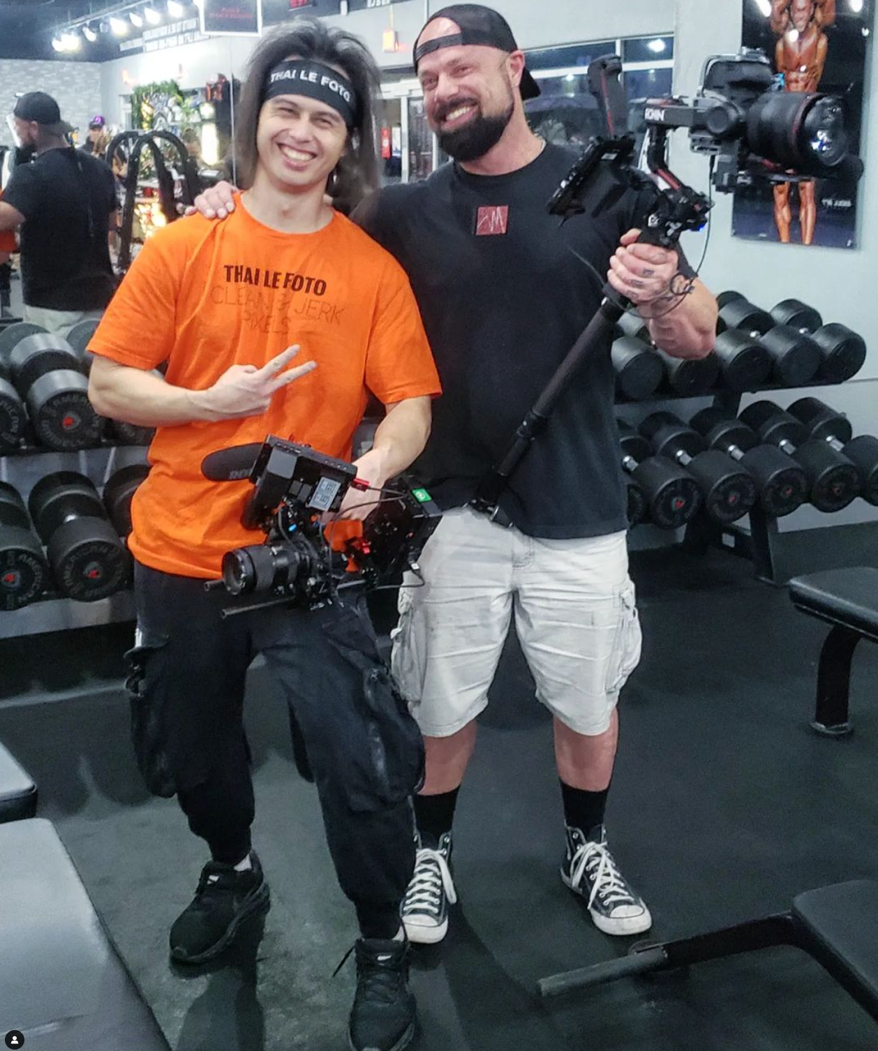 Two people posing with camera gear and smiling in a gym with weight racks visible in the background.