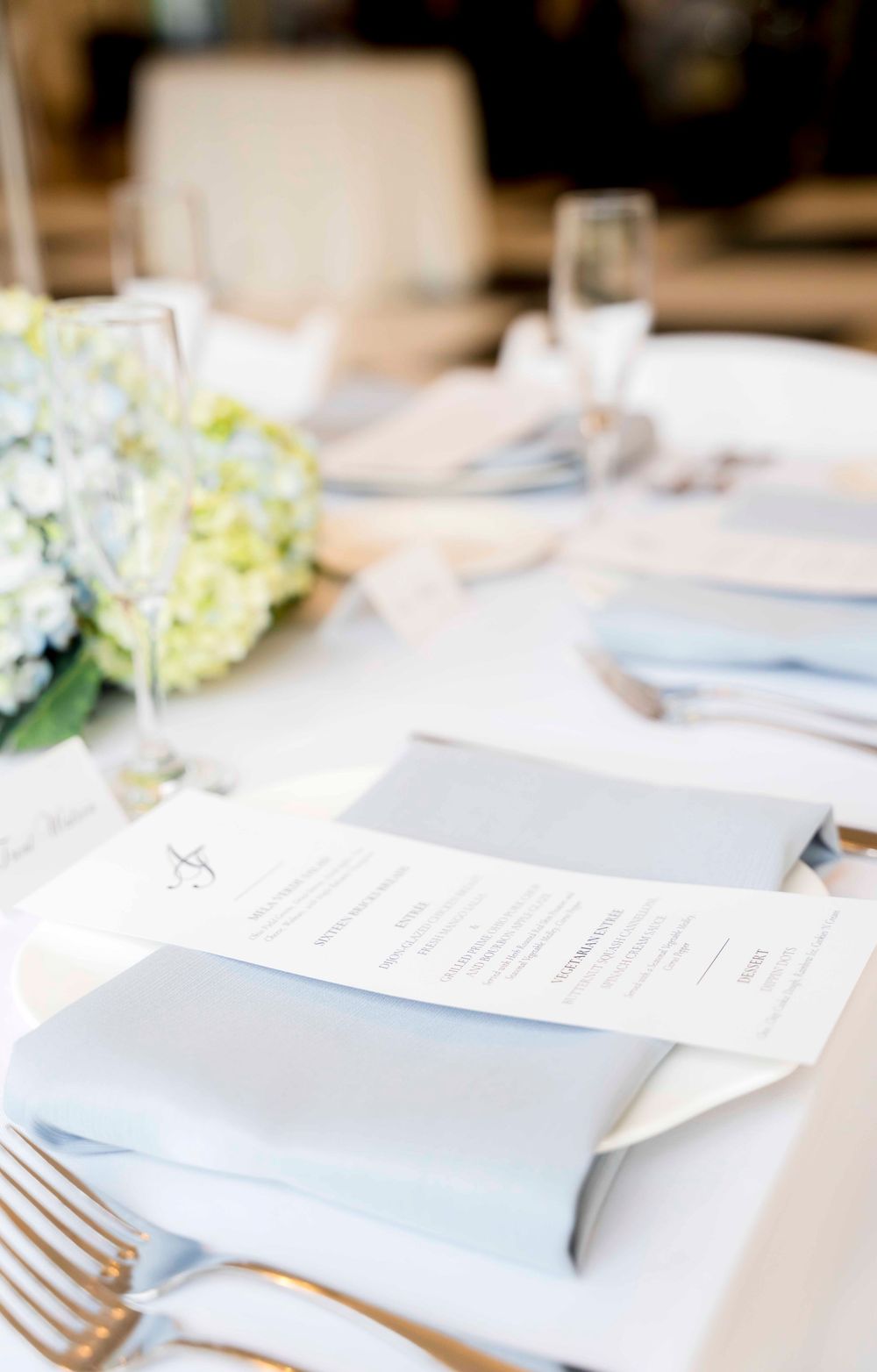 Elegant place settings at a wedding reception with white hydrangea centerpieces and menu cards on tables.
