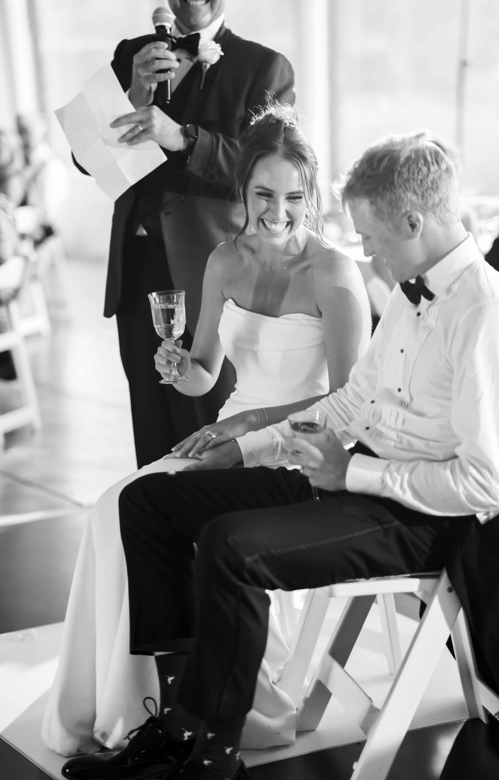 Wedding guests share joyful laughter during a candid black and white reception moment.