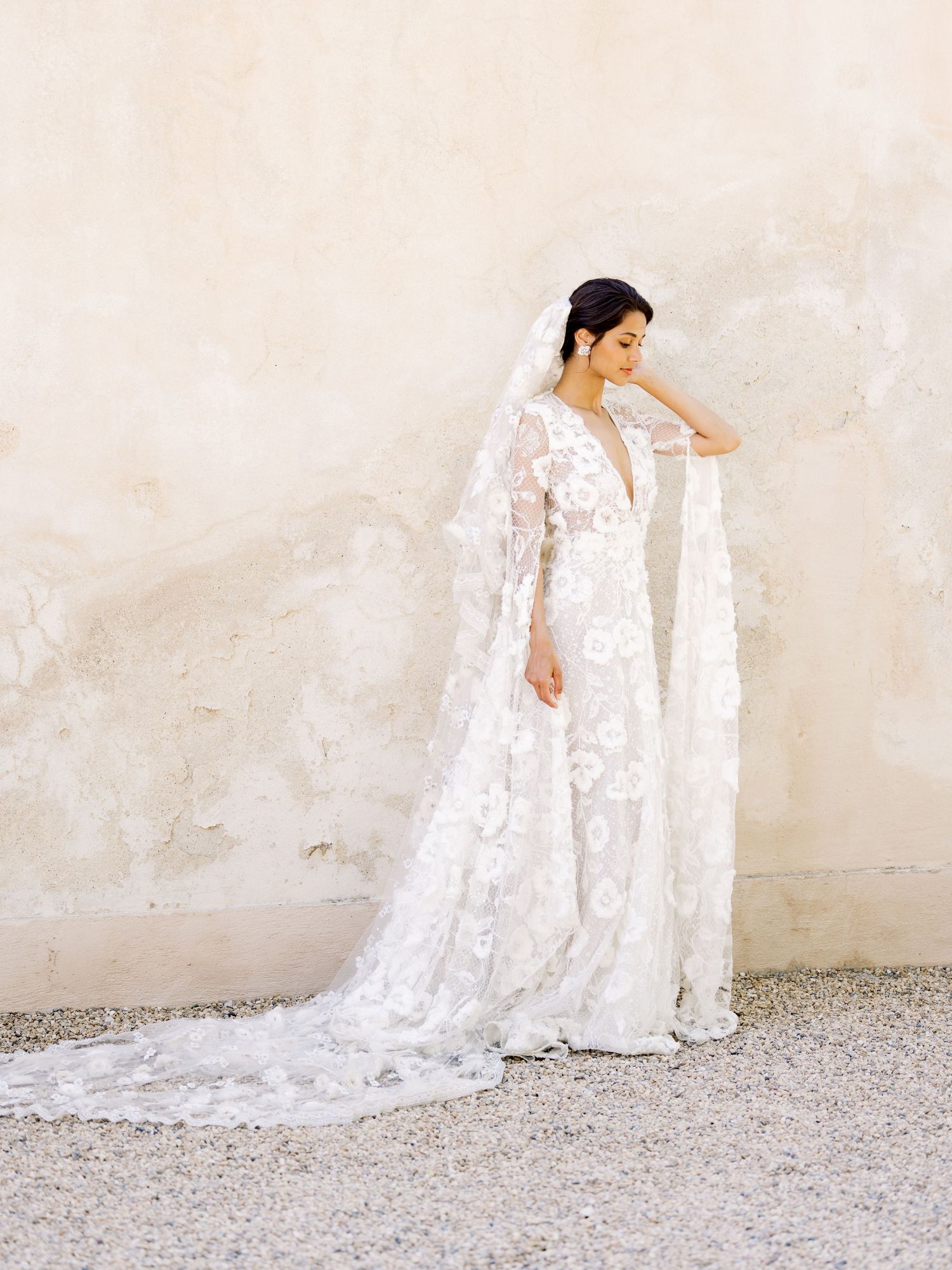 Ethereal bridal portrait featuring long-sleeved lace wedding gown with cathedral veil against neutral wall.
