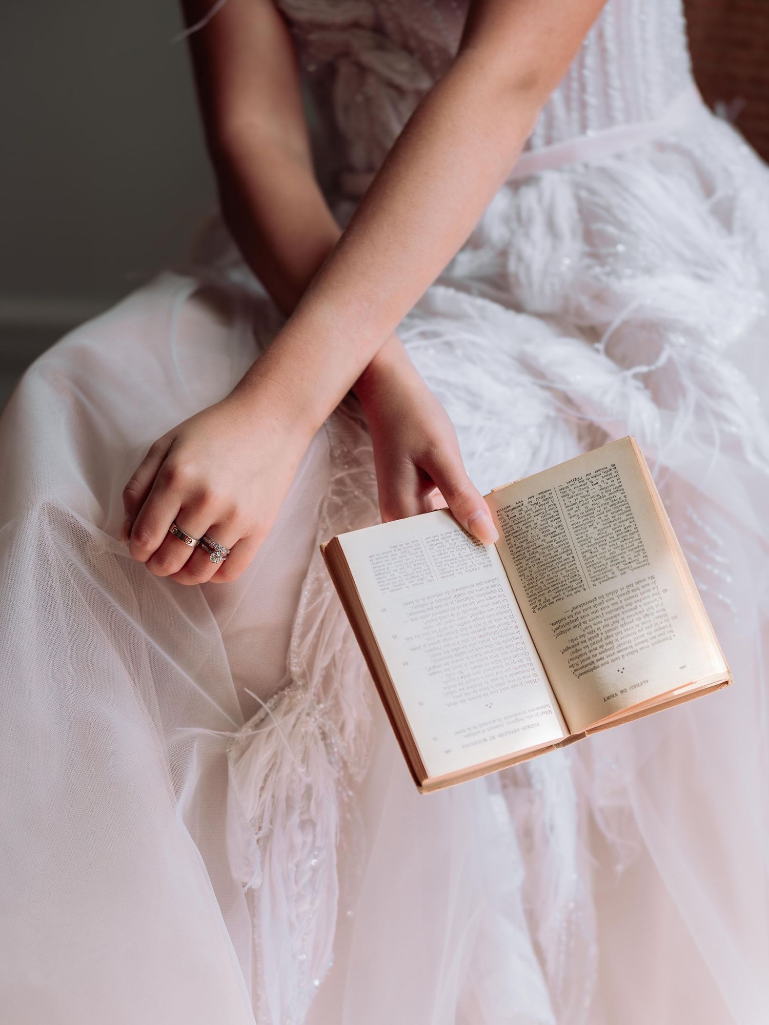 Hands holding an open book while wearing a white lace dress against soft bedding.