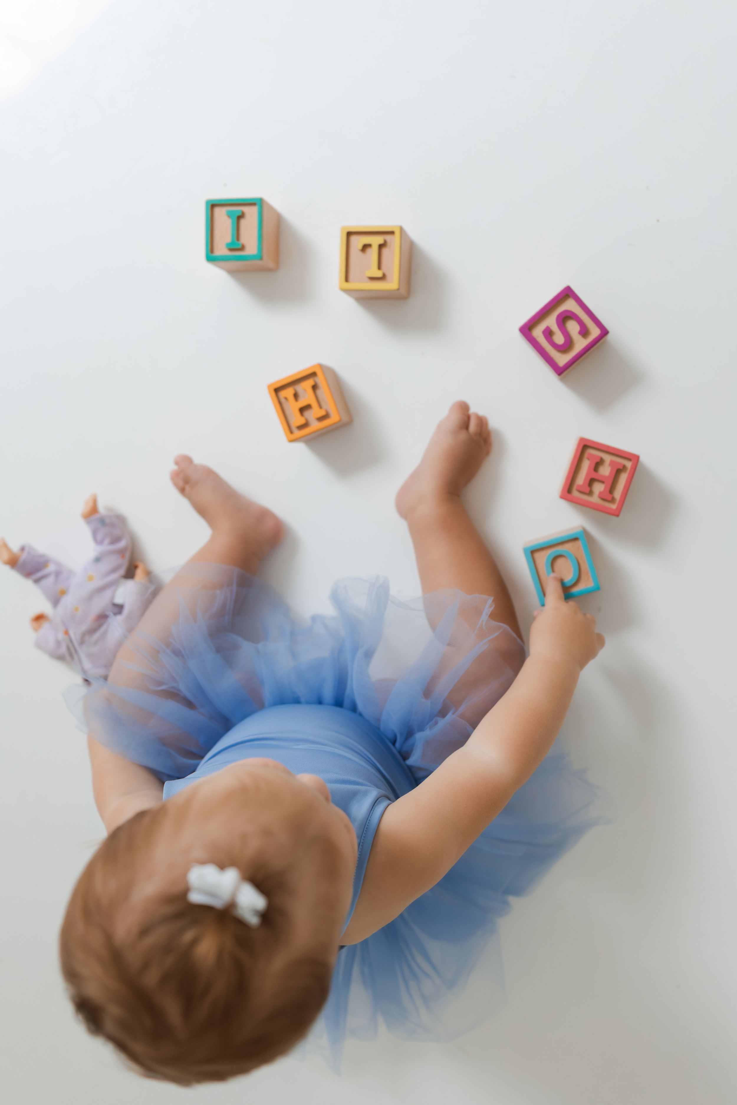 Baby in blue tutu dress lies on white background with colorful alphabet blocks scattered around.