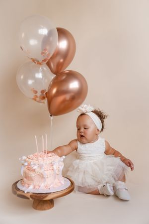 Studio photo series shows baby in white dress with first birthday cake and rose gold balloons against neutral backdrop.