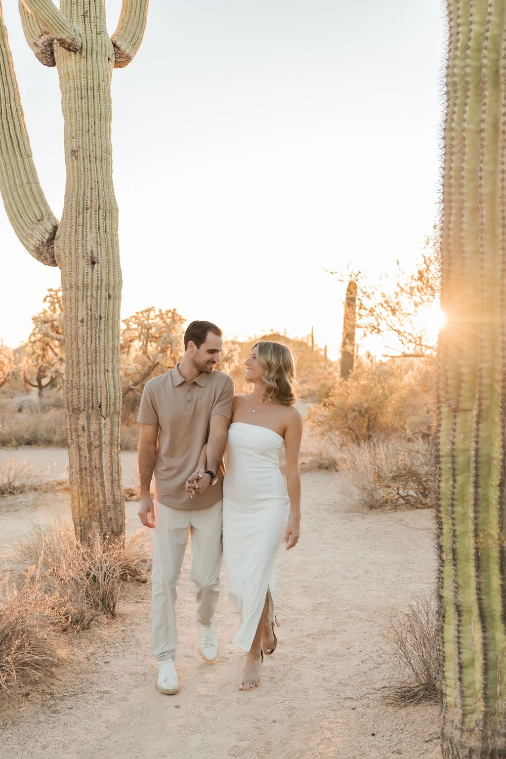 A couple walks together between tall saguaro cacti in the desert during golden hour sunset.
