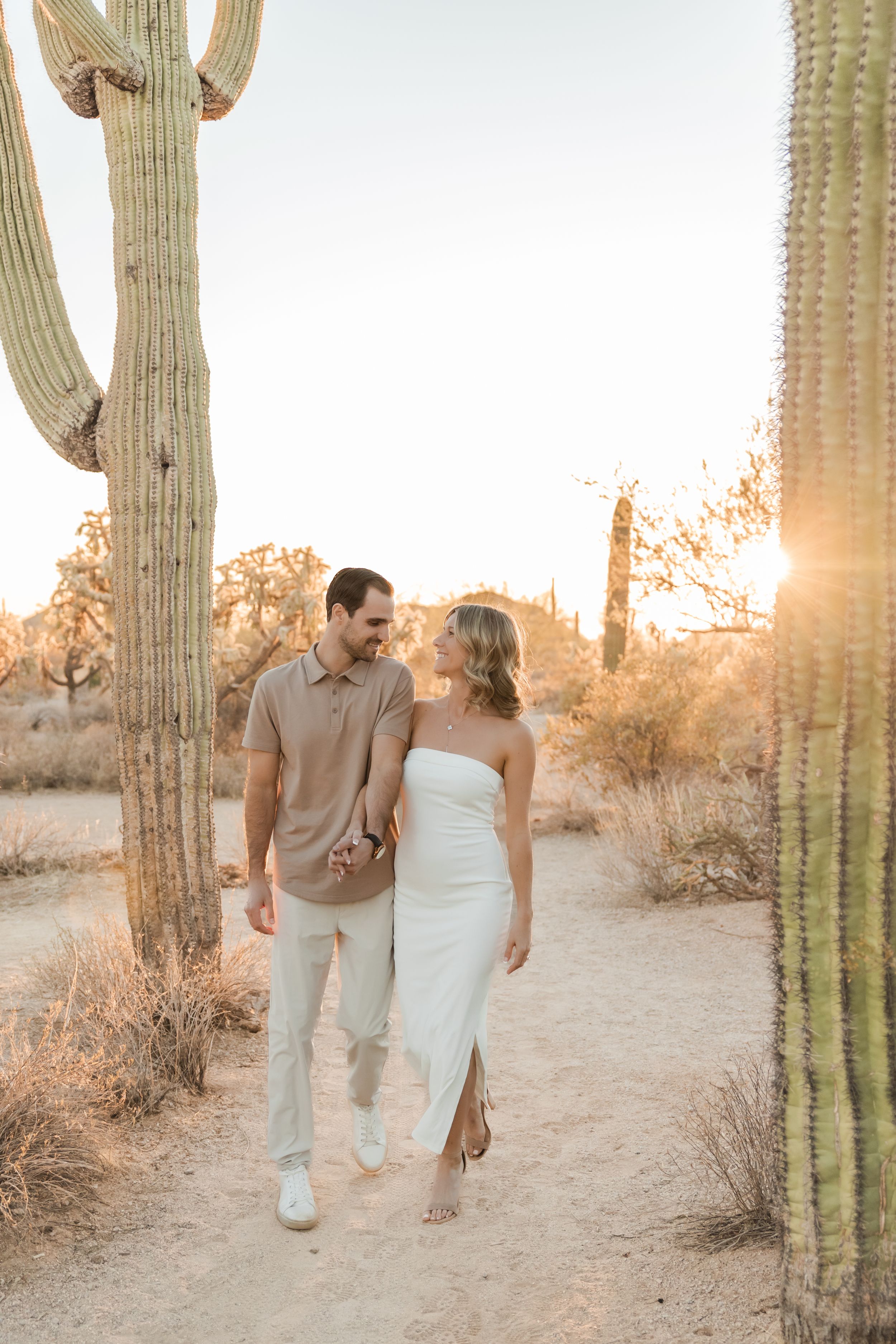 A couple walks together between tall saguaro cacti in the desert during golden hour sunset.