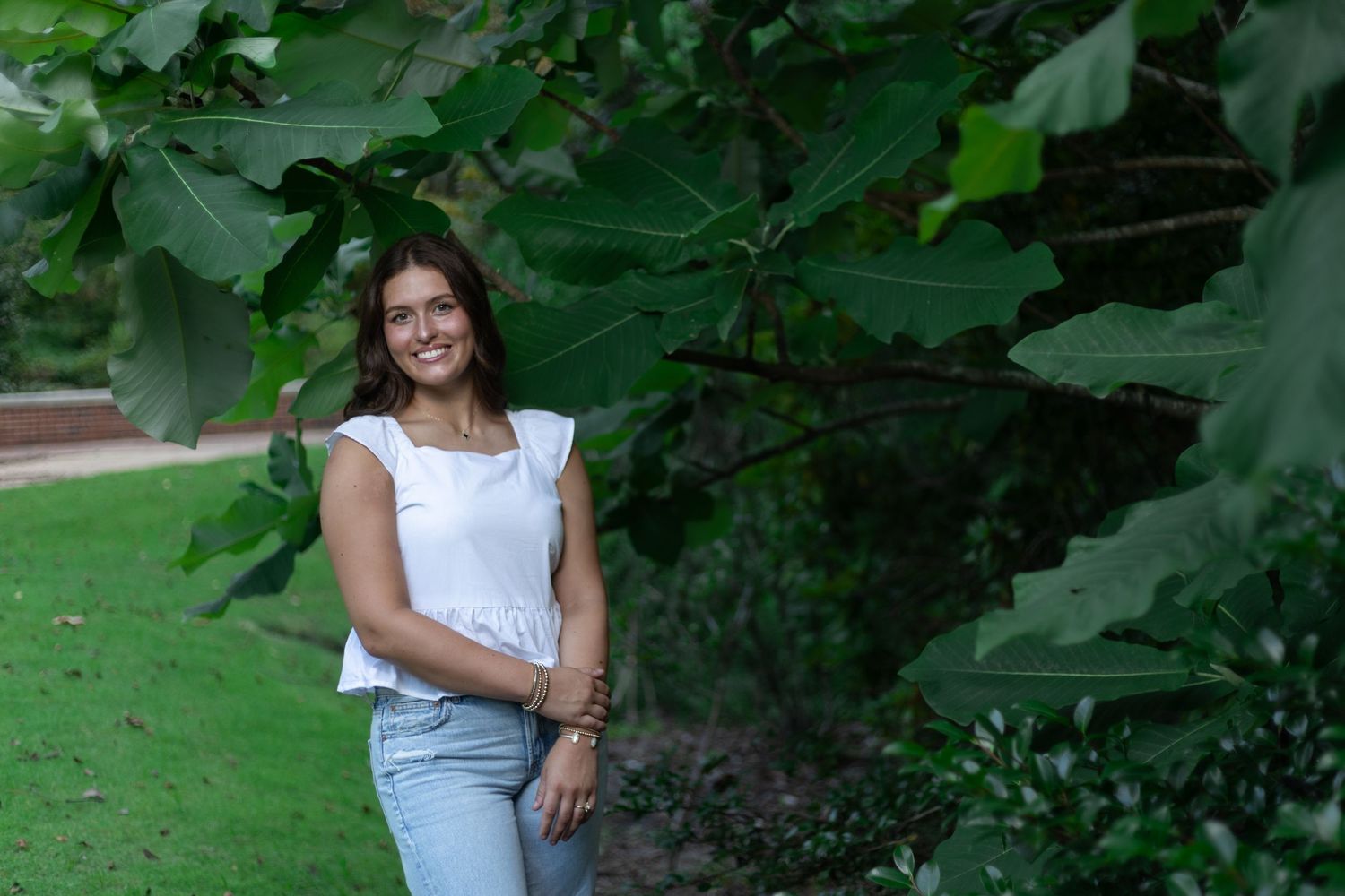 A senior poses in a lush green garden in Auburn Alabama wearing casual white shirts and denim.