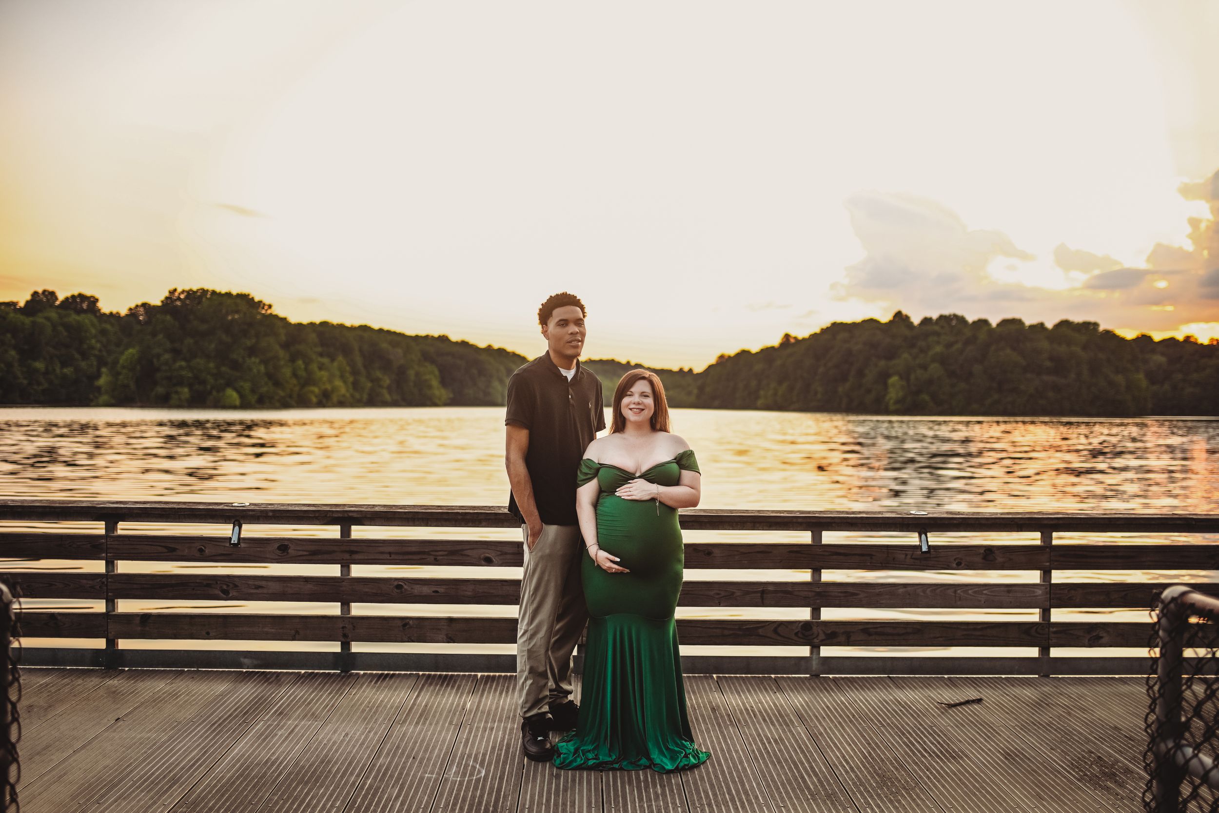 A couple poses on a wooden dock at sunset, with the woman wearing an elegant emerald green dress.