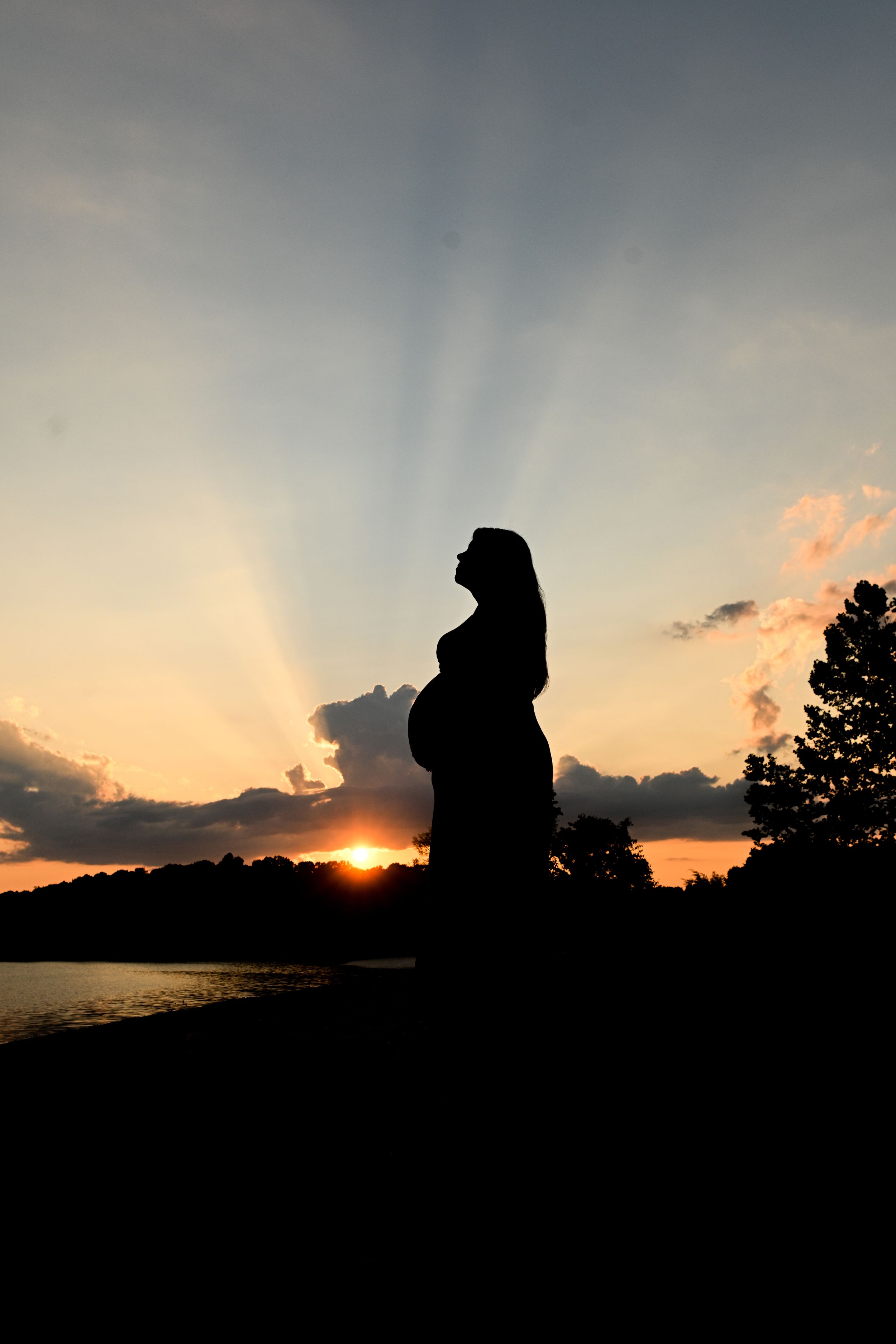 Silhouette of an expectant mother standing against a dramatic sunset over a lake.