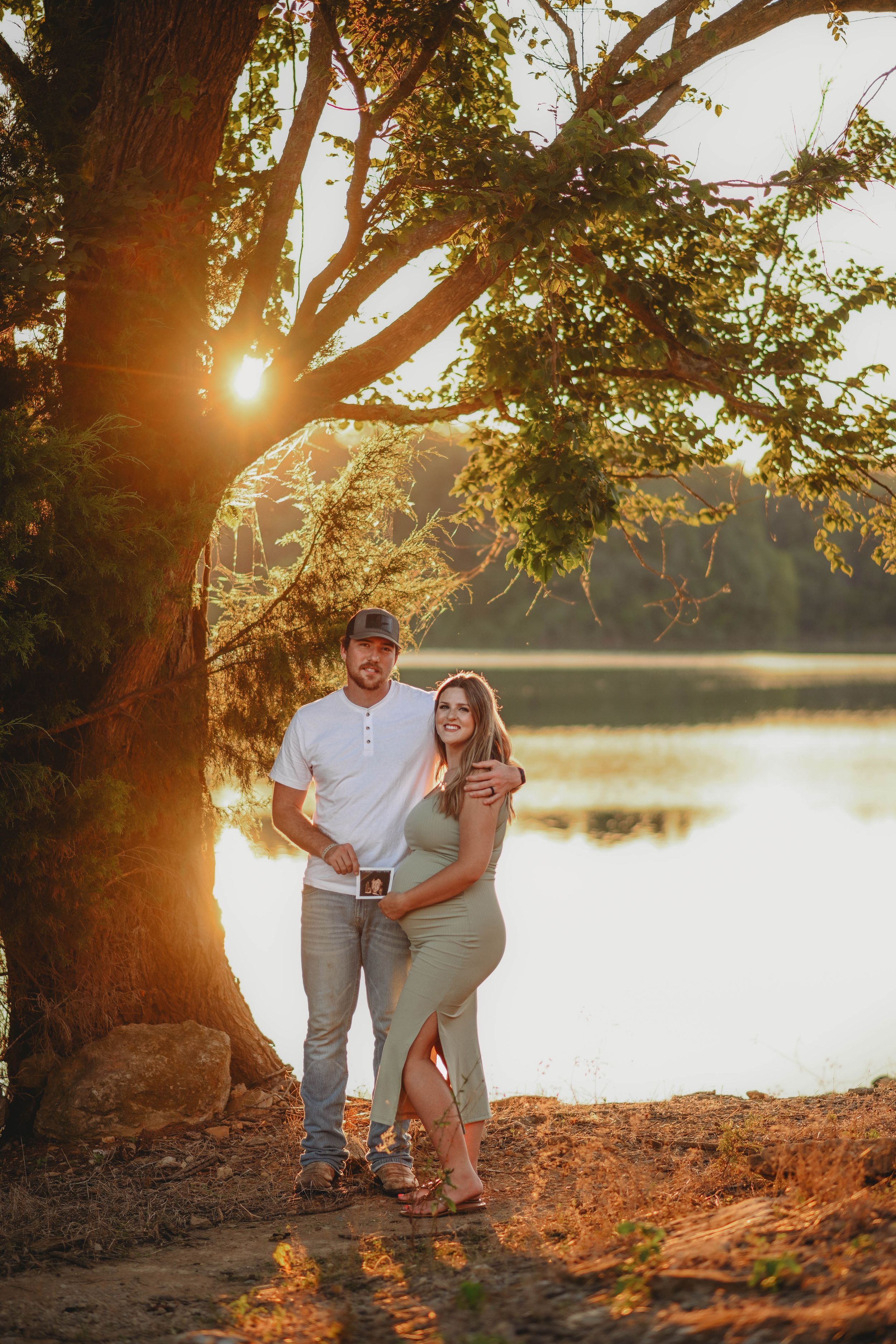 Couple poses by lake at sunset with sun flare through tree branches.
