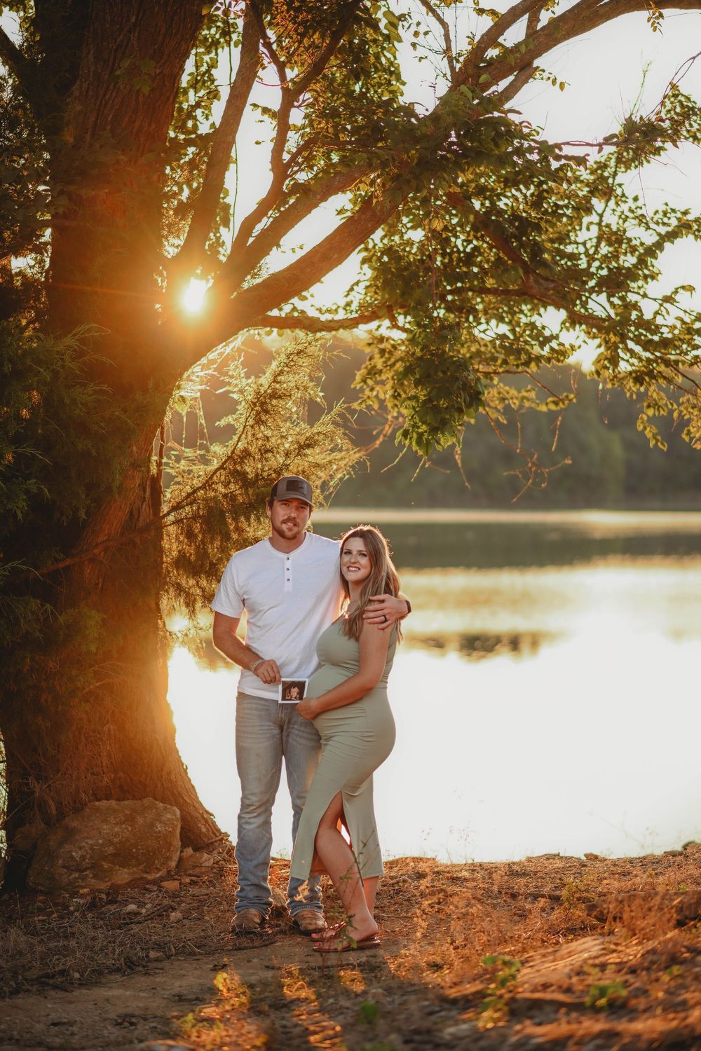 Couple poses by lake at sunset with sun flare through tree branches.