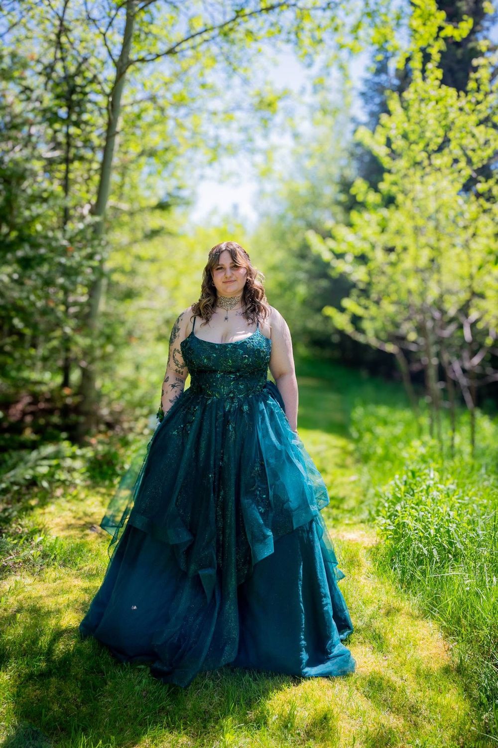 Young woman in elegant teal ballgown standing in a sunlit green forest during spring season.