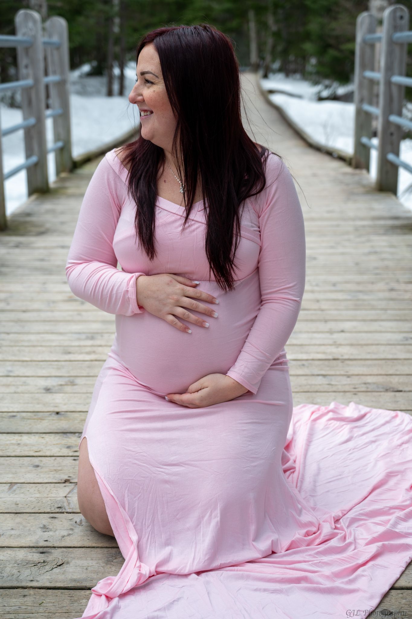 Smiling pregnant woman in pink maternity gown kneeling on wooden bridge surrounded by snowy forest.