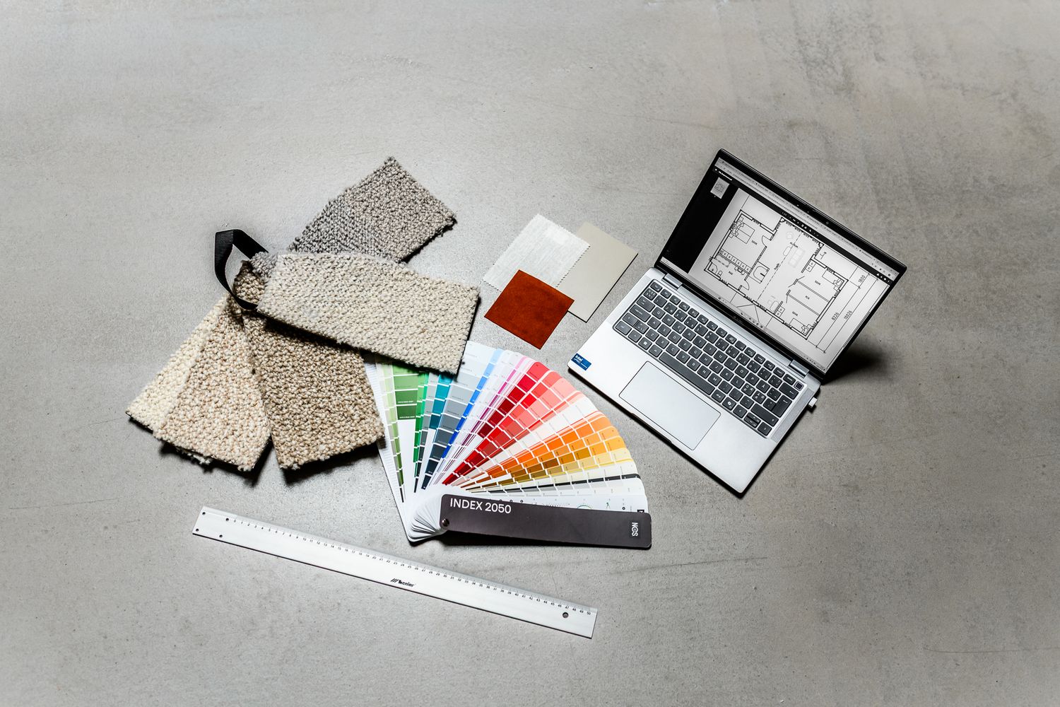 En dator, färgprover, mattprover, textilprover och en linjal ligger på ett golv. Flatlay för inredningsföretaget Studio Karlsson i Falköping