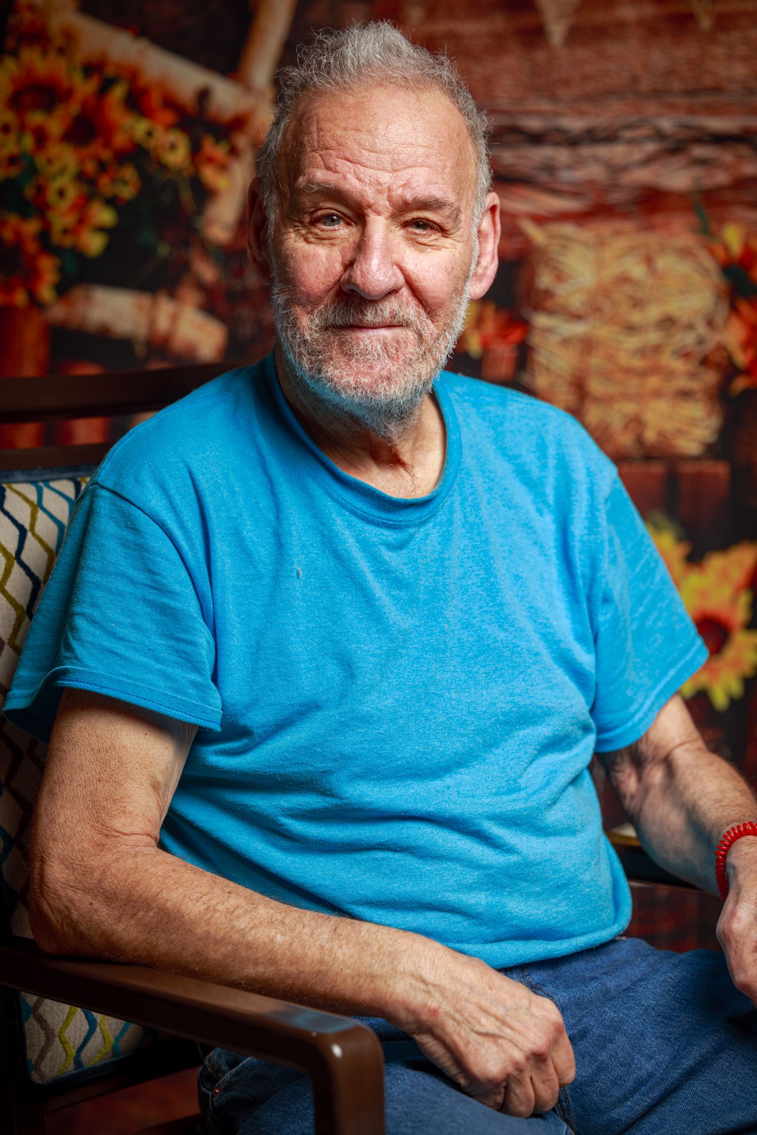 Smiling senior in bright blue t-shirt sits comfortably in wooden chair against rustic brick wall with autumn decor.