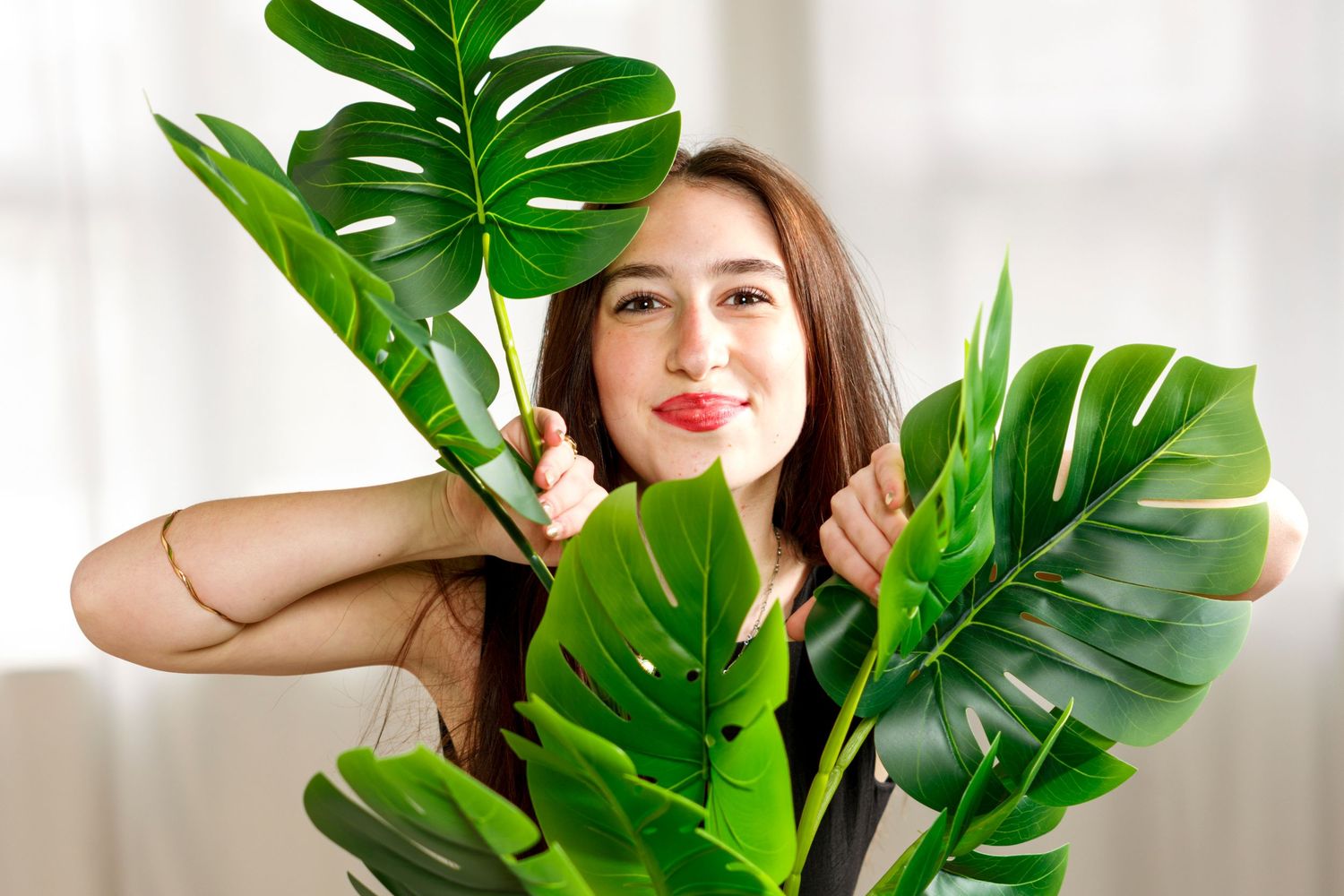 A person poses playfully with large monstera delicata leaves against a bright white background.