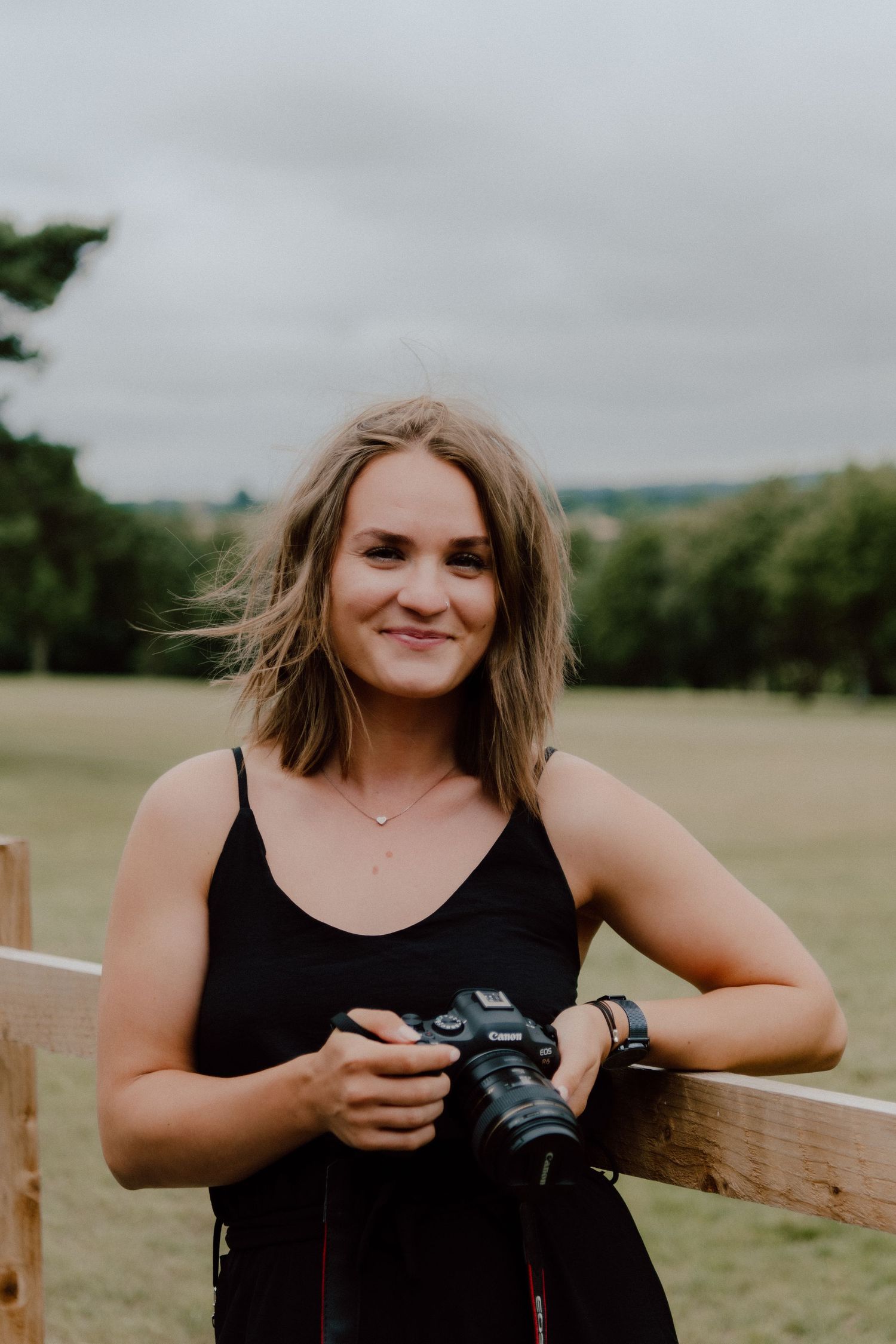 A smiling photographer in a black tank top holding a camera outdoors.