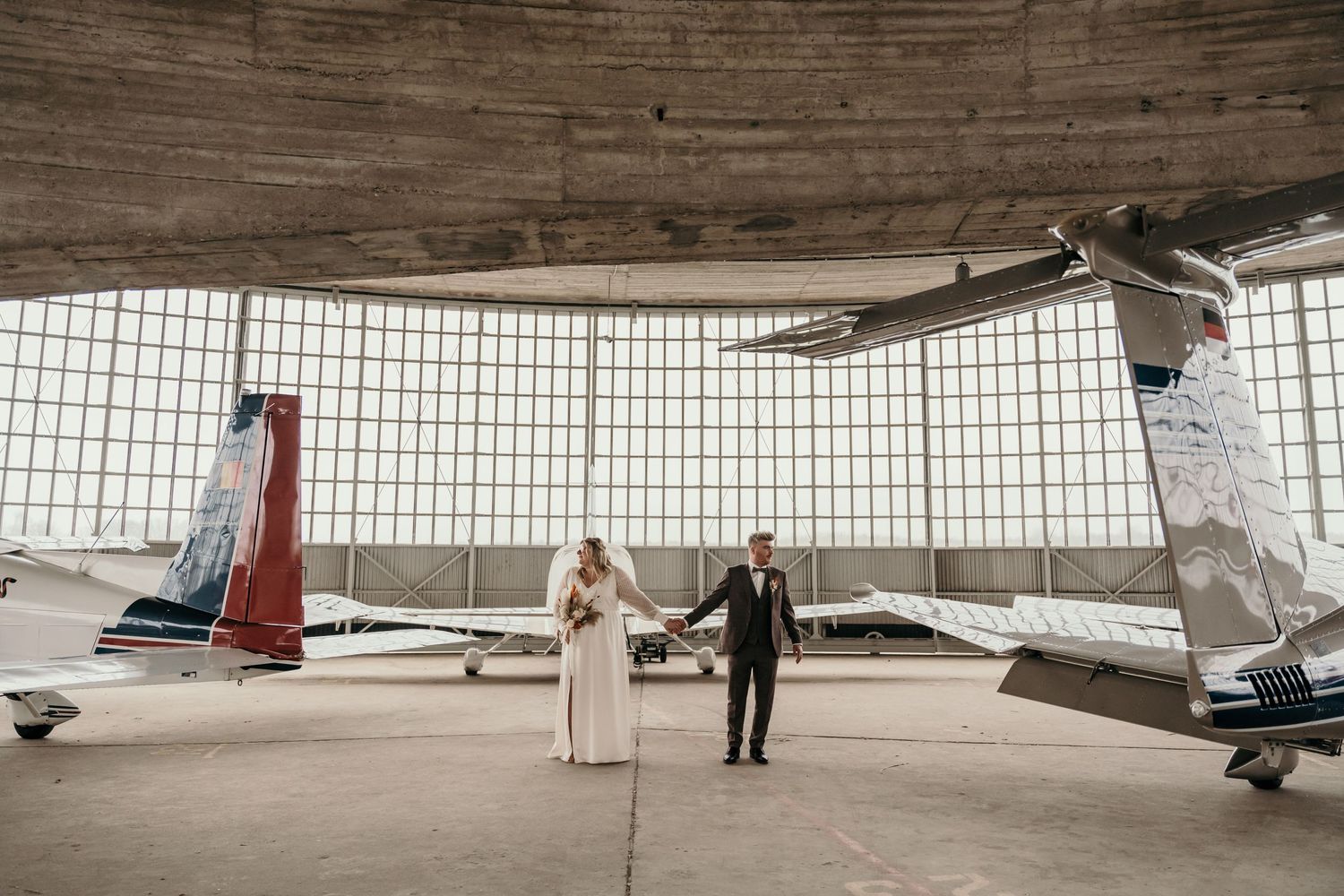Een getrouwd koppel, hand in hand, op een mooie fotolocatie in Grimbergen, het vliegveld. De sfeer van de industriële loods geeft veel sfeer