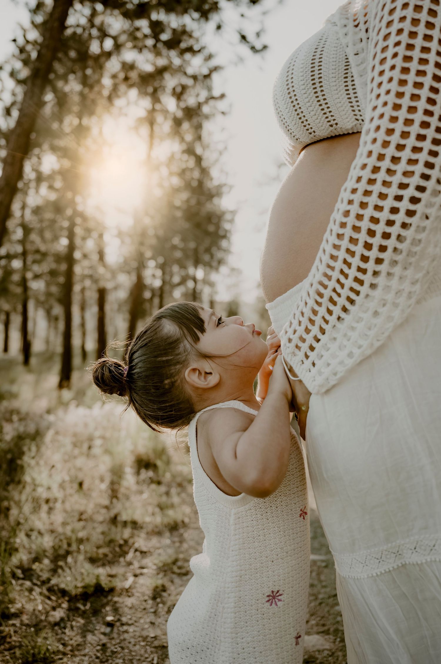 Intimate maternity moment with child kissing pregnant belly in sunlit forest.