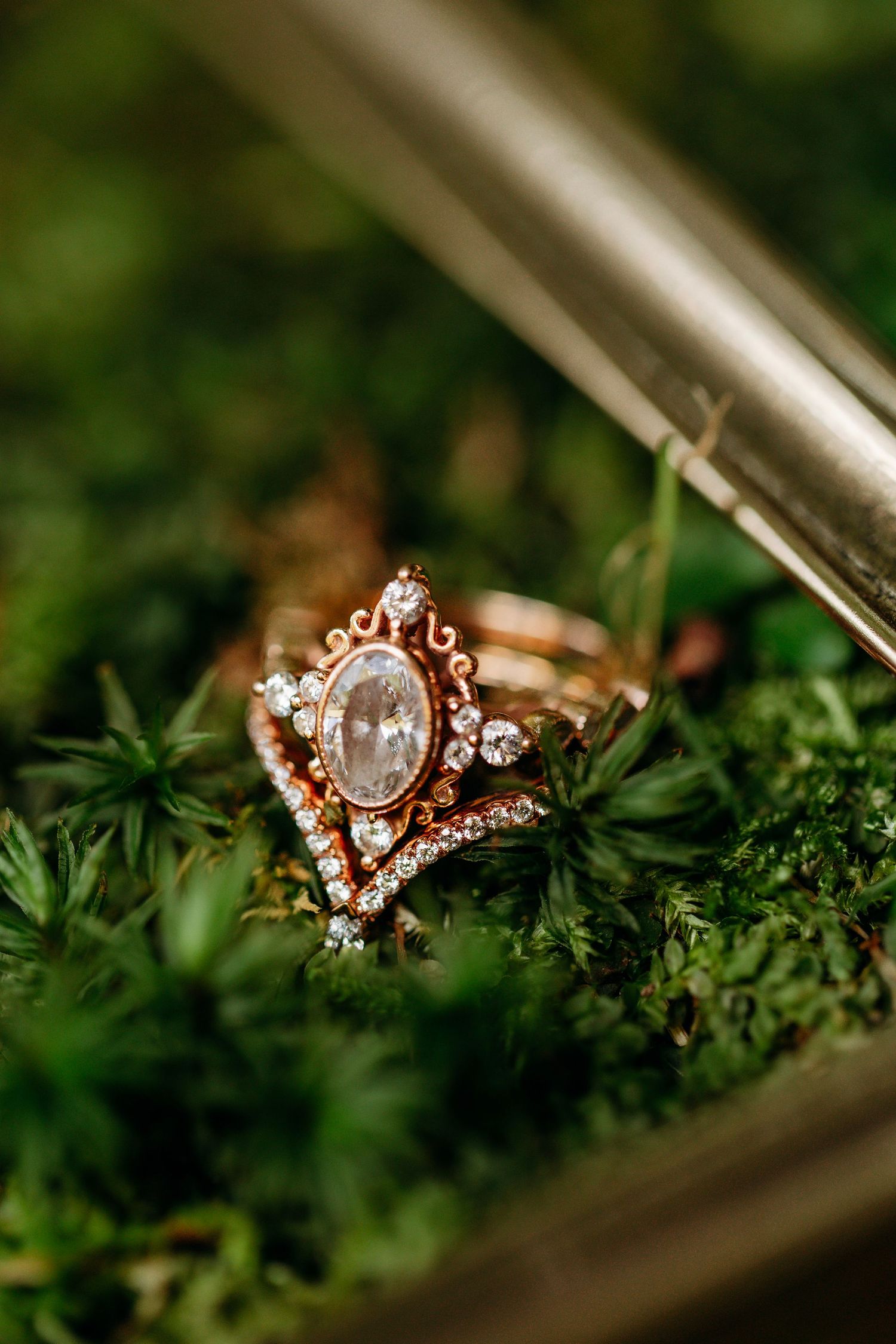 An ornate vintage-style rose gold engagement ring with oval stone rests on green moss.