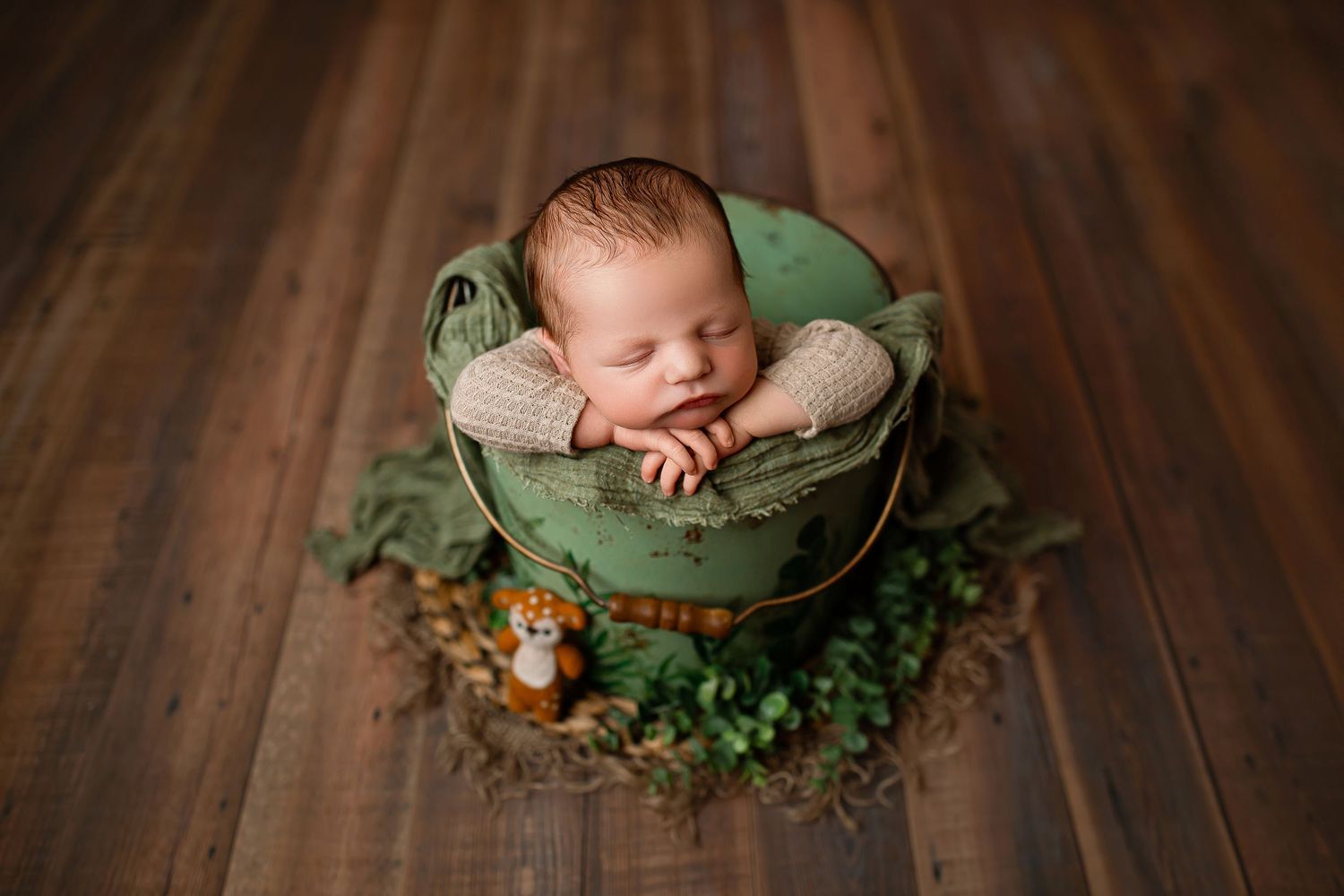 Newborn baby boy sleeping in vintage green bucket with sage wrap and greenery on rustic wood floor at Flowertown Photography Summerville SC