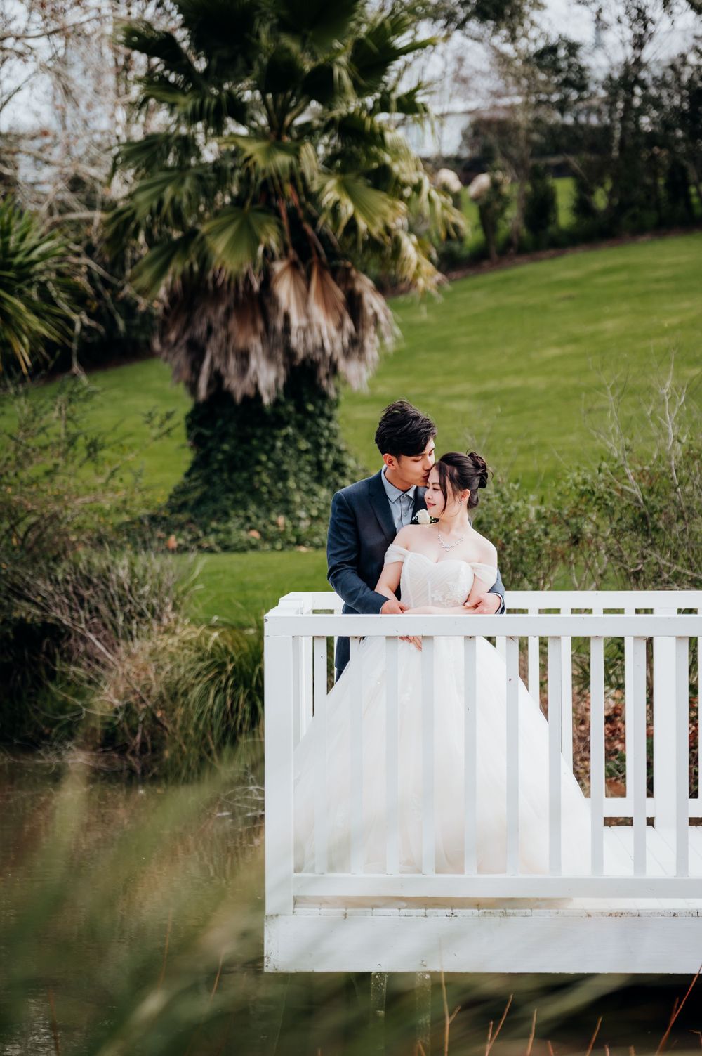Newlyweds share an intimate moment on a white bridge surrounded by palm trees.