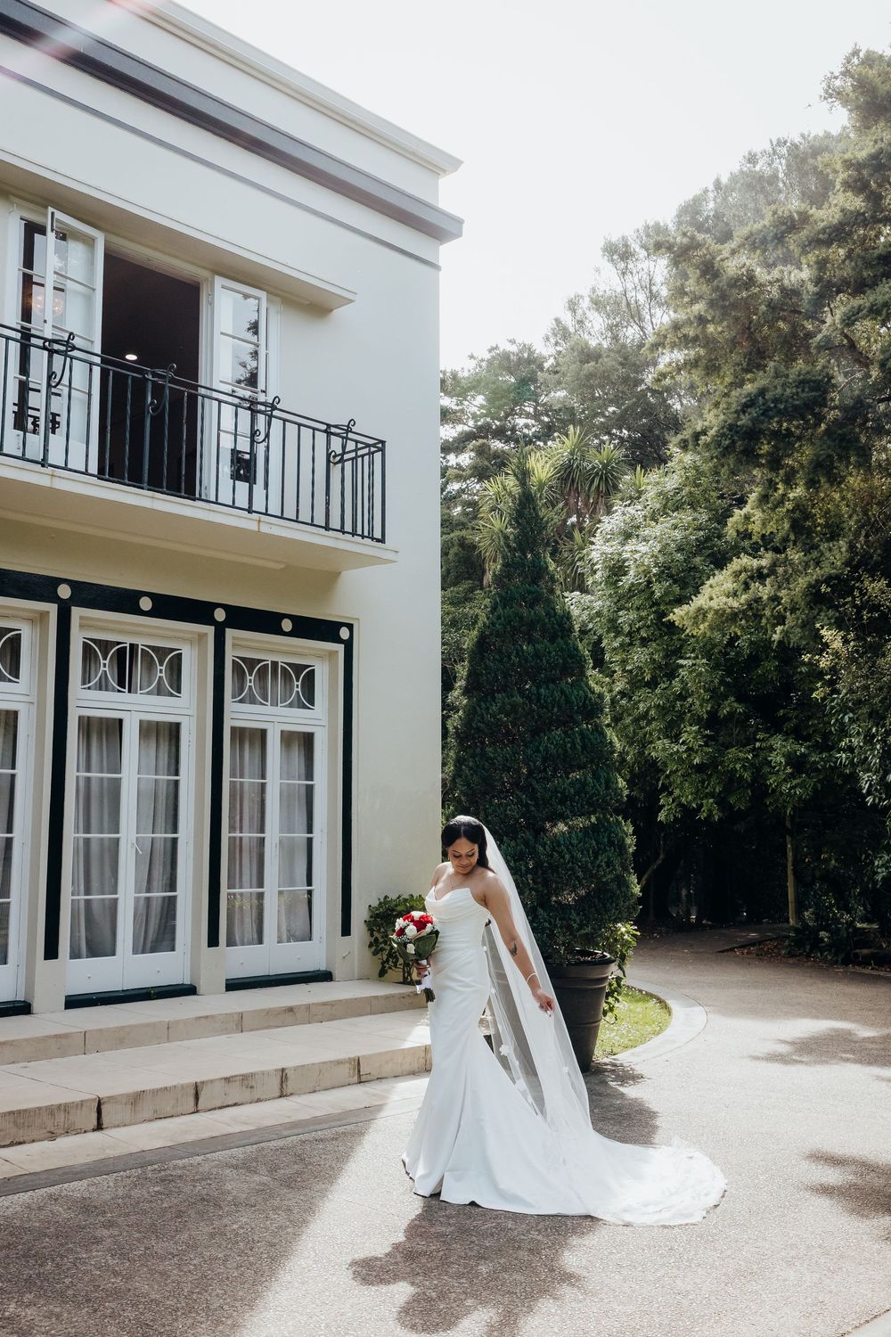 Bride in white gown and veil holds bouquet outside elegant two-story venue with balcony and lush trees.