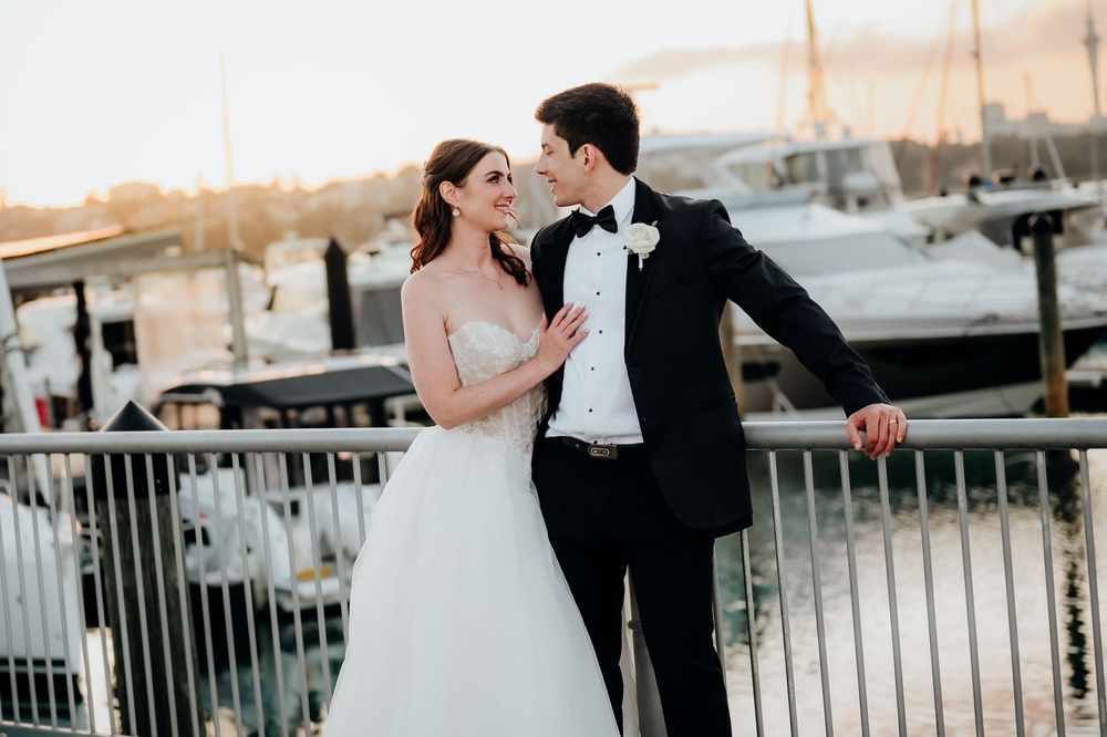 Bride and groom share a romantic moment on a marina dock at sunset, with sailboats in the background.