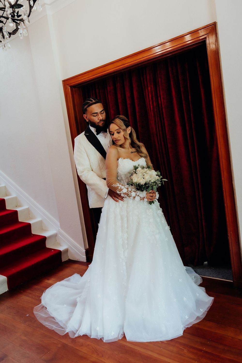 Bride in white ball gown holding bouquet stands with groom in white tuxedo jacket near red curtains and staircase.