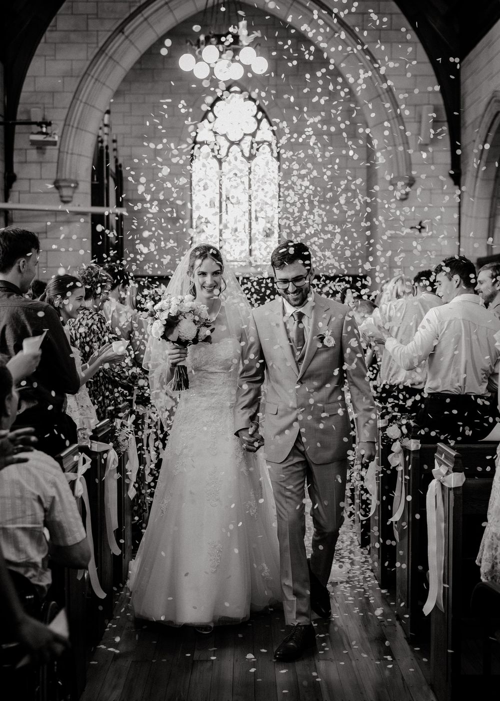 Black and white photo of a bride and groom walking down the church aisle as guests throw confetti.
