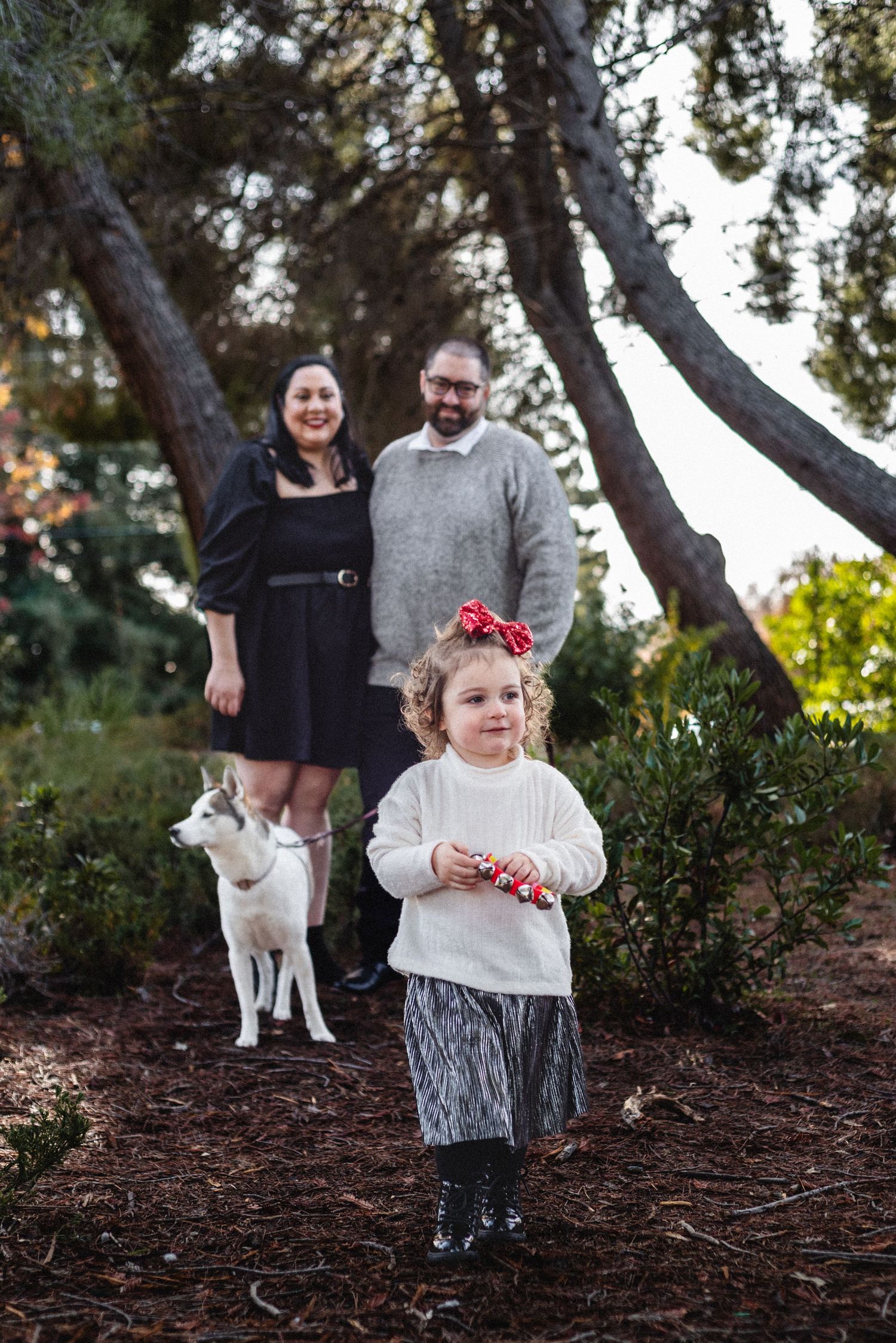 Family portrait in a wooded area with a white dog and child wearing a festive hair accessory.