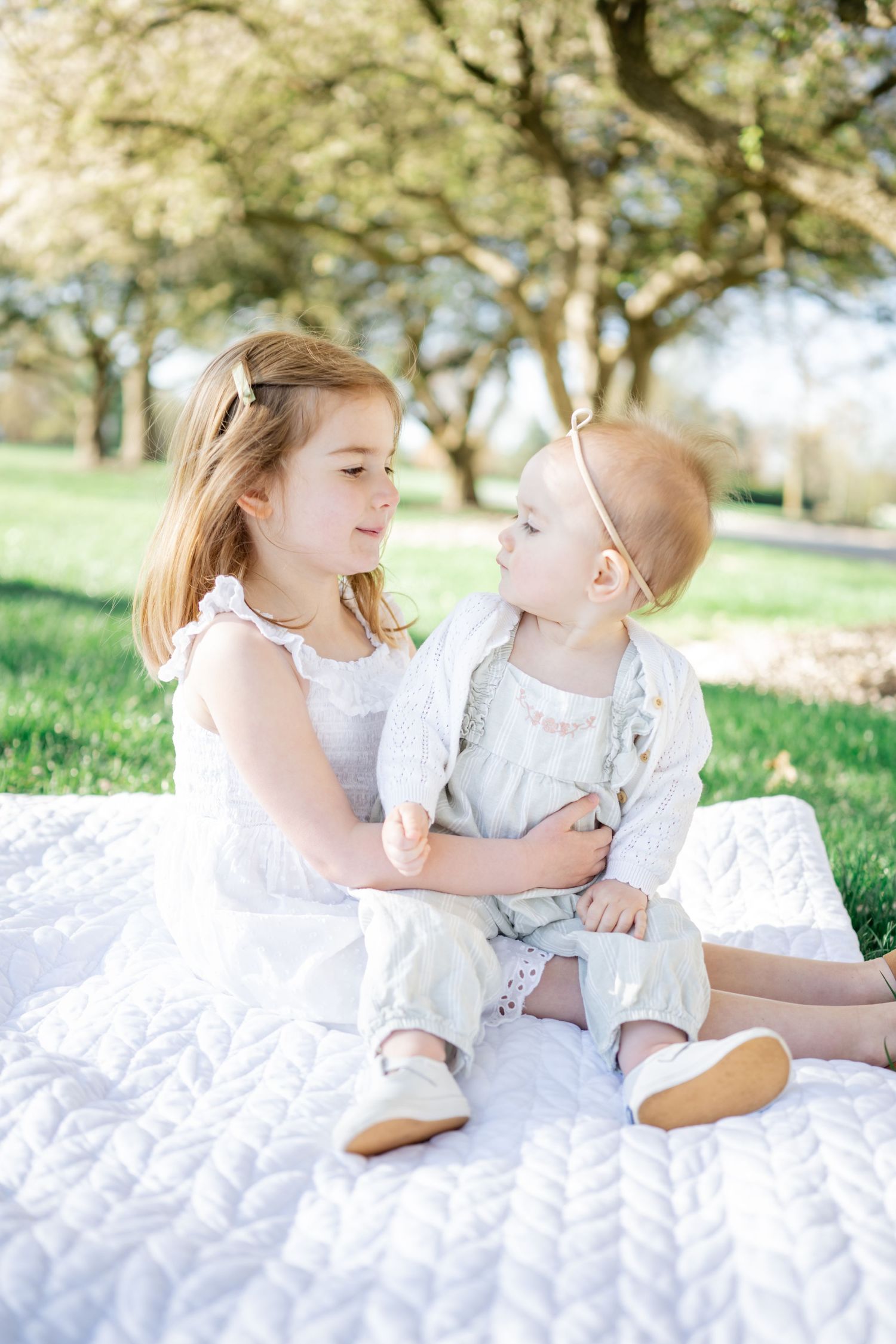 indianapolis family photographer, sisters smile at each other while sitting on white blanket at park