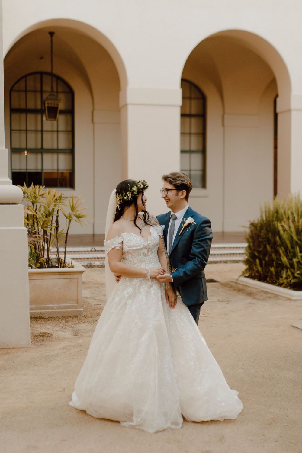 A wedding couple stands before arched windows in warm afternoon light.