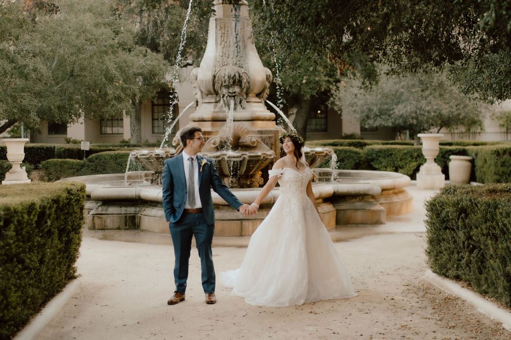 A wedding couple poses by an elegant stone fountain in a manicured garden setting.