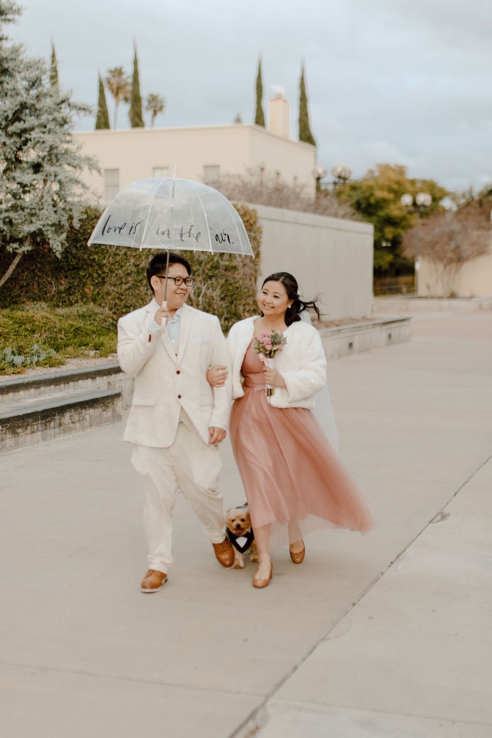 A couple in vintage-inspired outfits walks with a clear umbrella and teddy bear on a cloudy day.