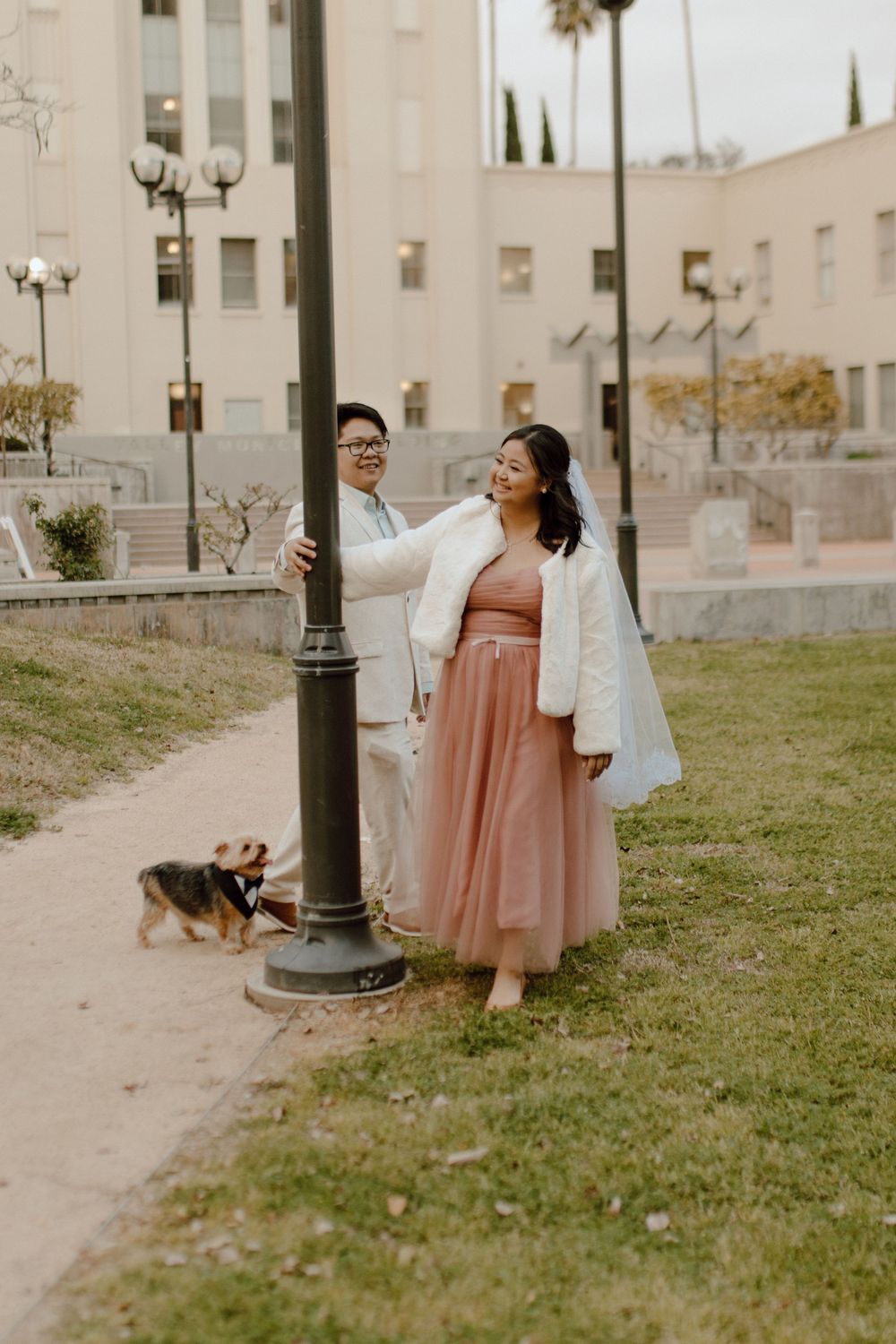 Couple in formal wear poses by vintage lamppost with small dog in urban park setting at sunset.