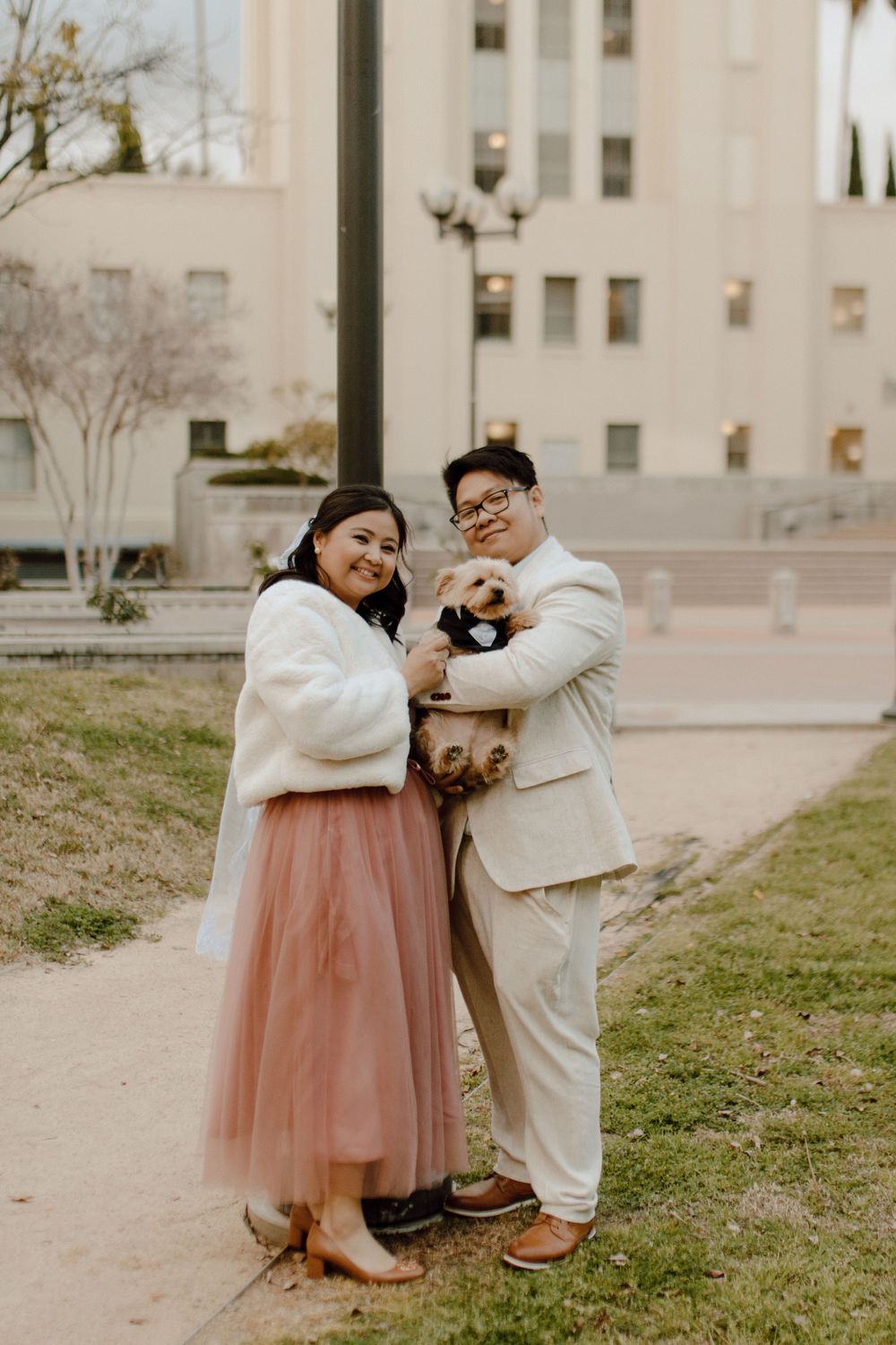 Couple in white and beige formal wear embraces while holding small dog at city park.