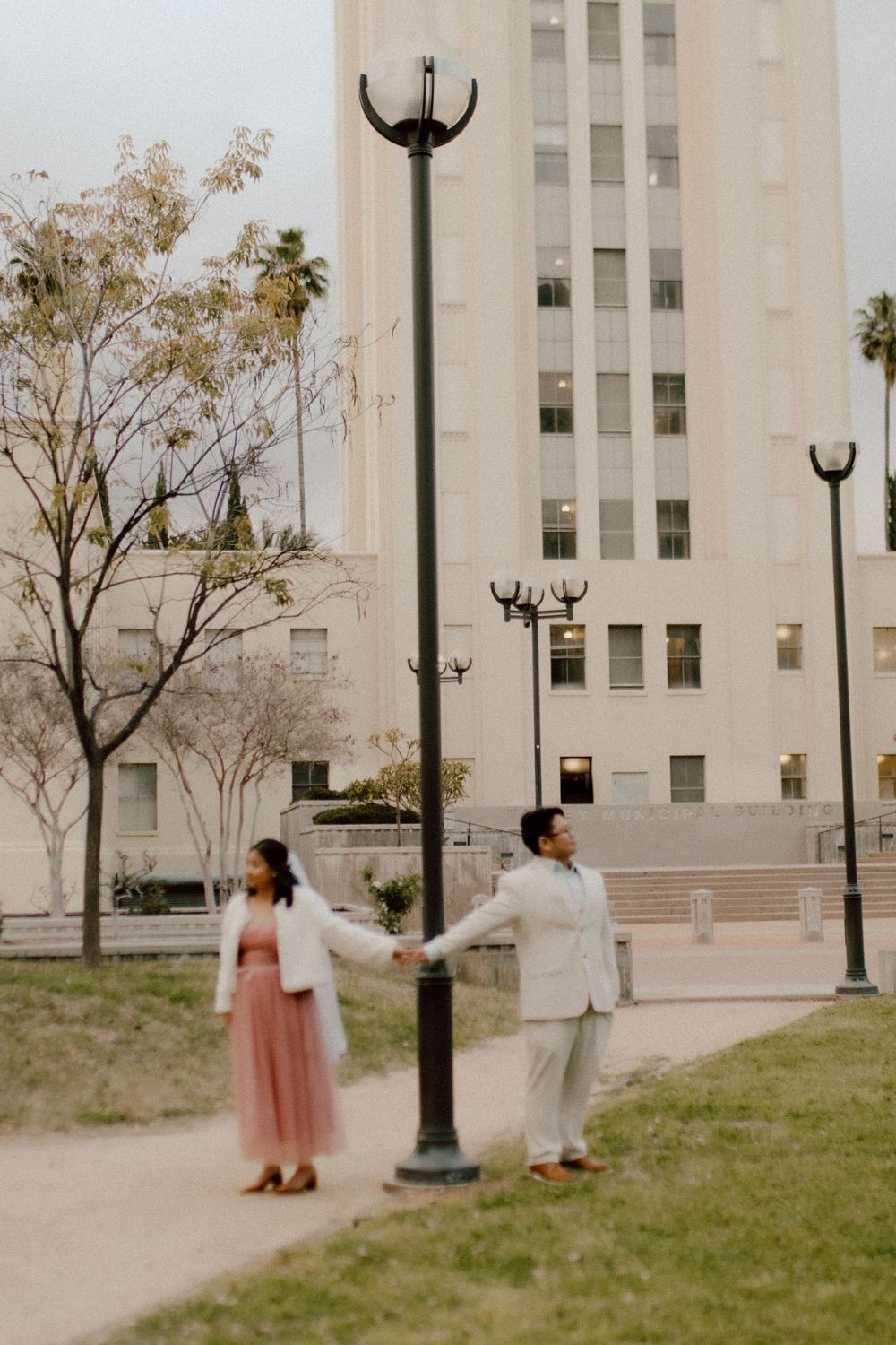 Couple in formal attire poses playfully with lamppost in urban garden setting.