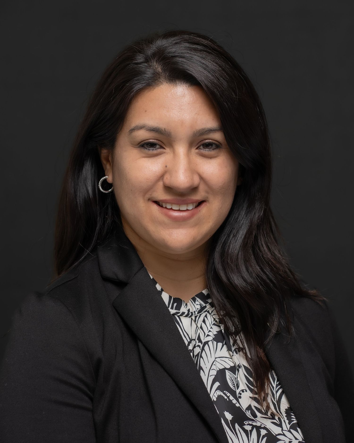 Professional headshot of smiling woman in black blazer and patterned blouse, posed against a dark studio background